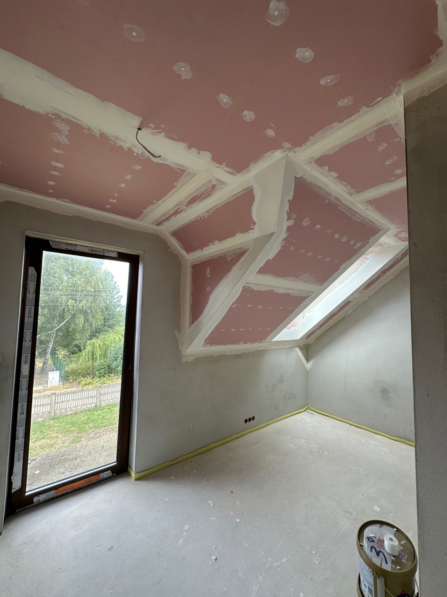 Pokój na poddaszu w trakcie prac wykończeniowych: płyty karton-gips na suficie, szpachlowane łączenia, surowa podłoga, okno z widokiem na zieleń, wiadro z farbą w rogu.