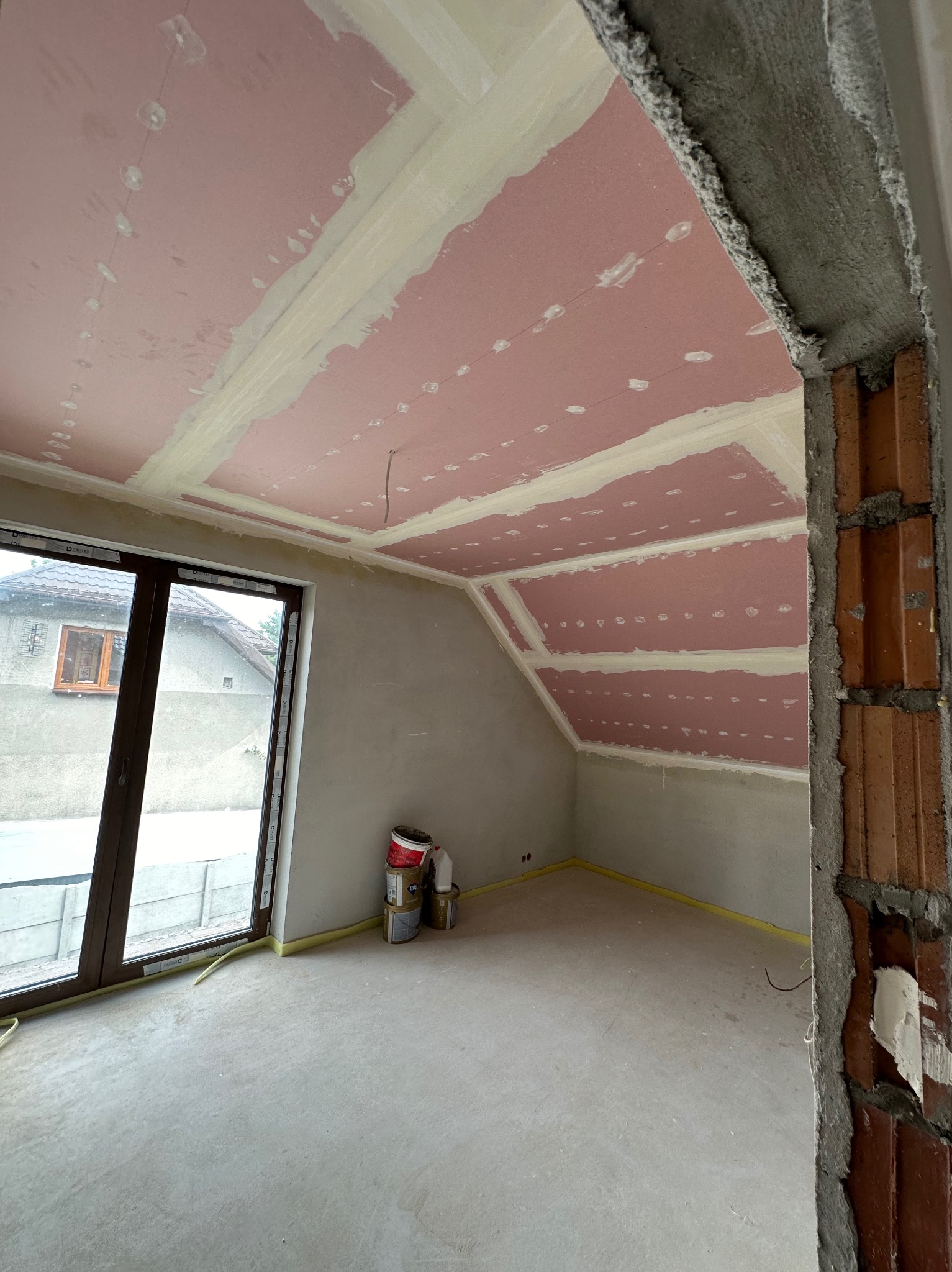 Poddasze w trakcie prac wykończeniowych: różowa płyta karton-gips na skosach sufitu, szpachlowane łączenia, surowy betonowy filar, okno z widokiem na dom.
