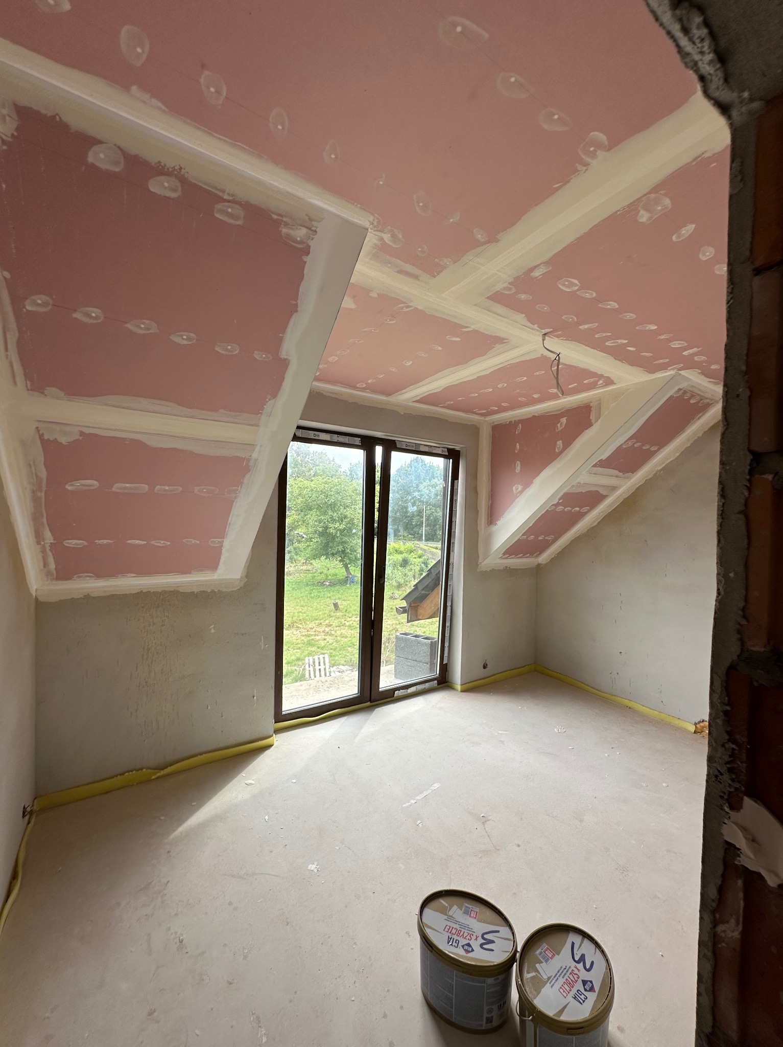 Poddasze po montażu płyt gipsowo-kartonowych, z widocznymi łączeniami i szpachlowaniem. Dwa wiadra z masą szpachlową na podłodze. Okno z widokiem na zieleń.