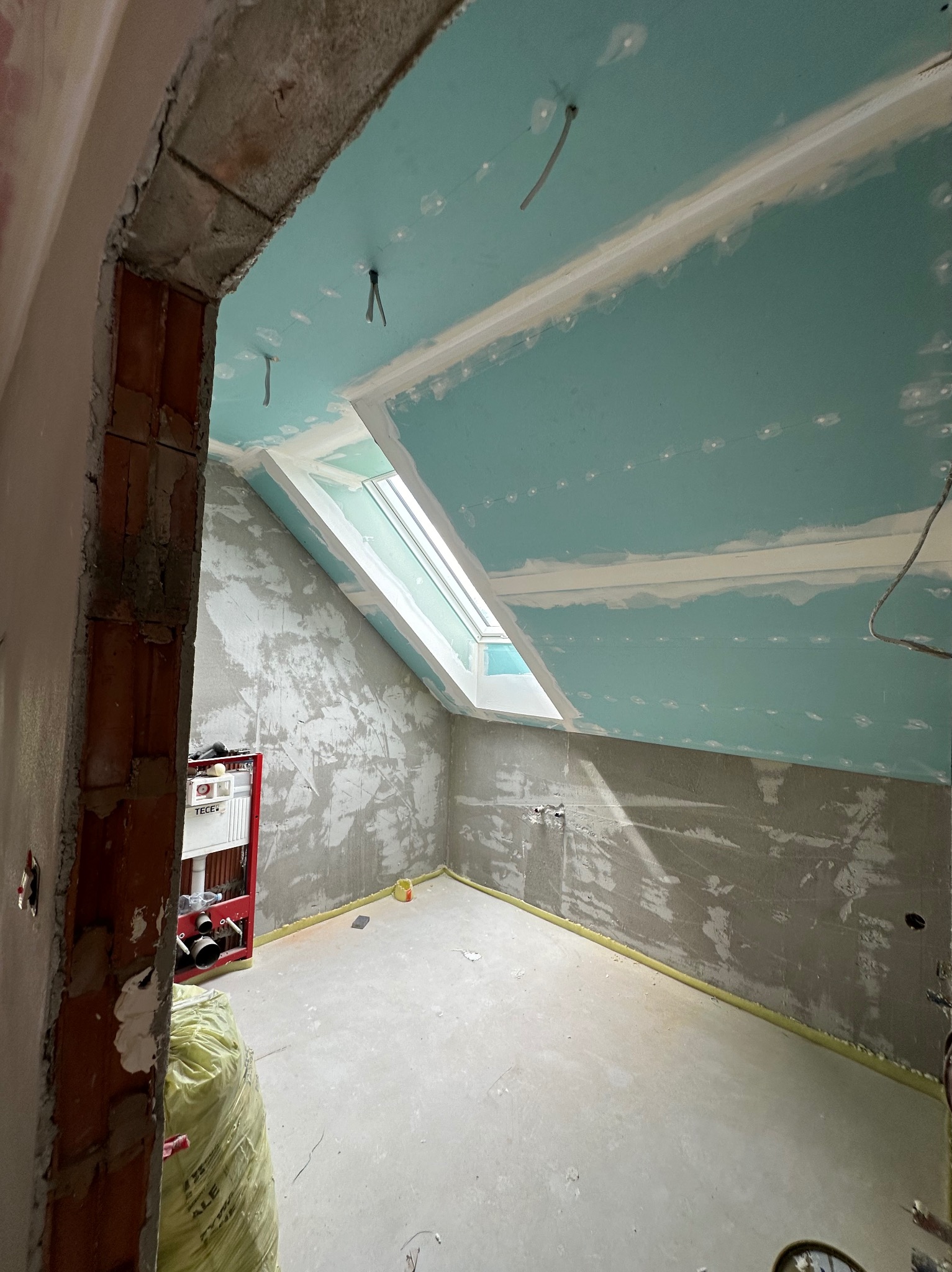 Pokój na poddaszu w trakcie prac wykończeniowych: ściany wyszpachlowane, sufit z płyt karton-gips, okno dachowe. Widoczny stelaż podtynkowy WC i worek z materiałami budowlanymi.