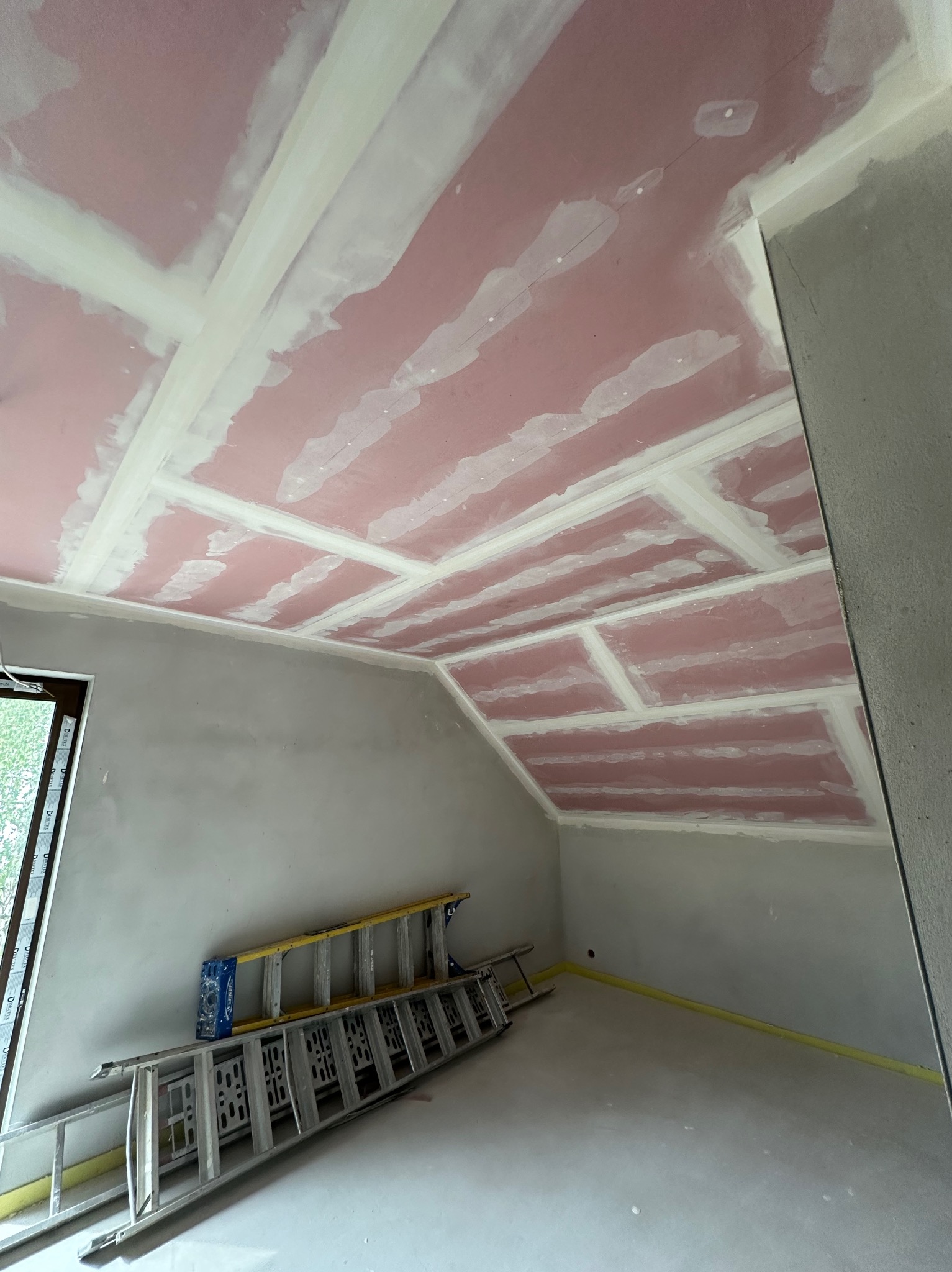 Poddasze z płytami karton-gips na suficie, przygotowane do szpachlowania. Widoczne łączenia płyt i drabina oparta o ścianę. Taśma malarska na podłodze.