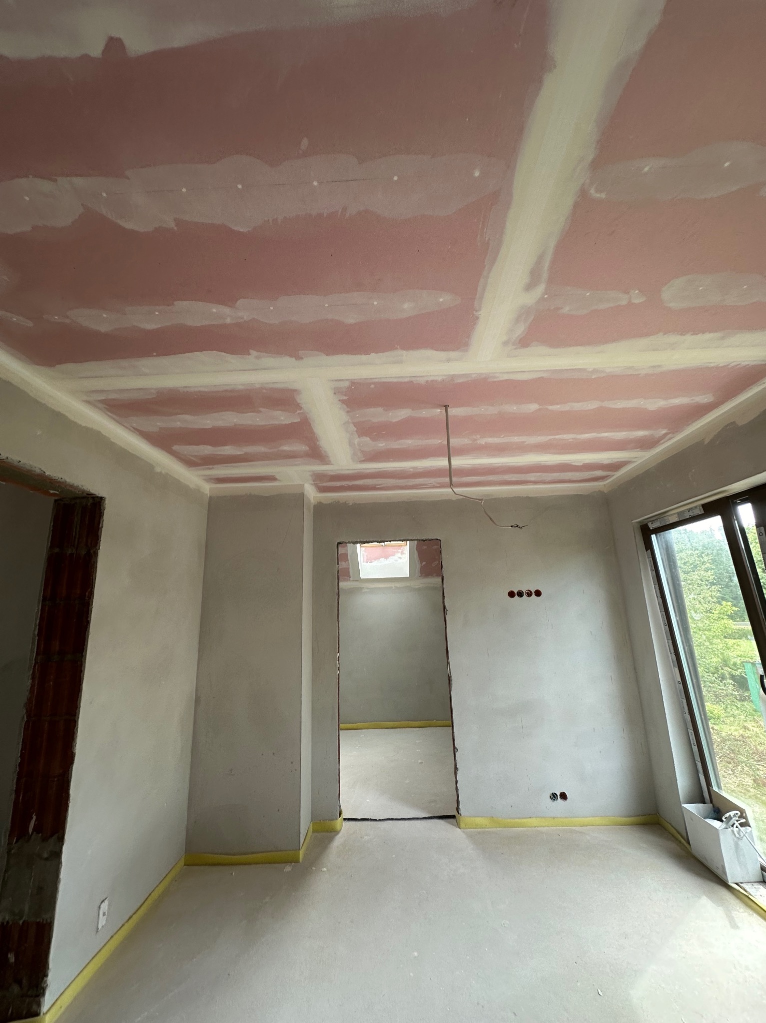 Pokój w trakcie prac wykończeniowych: ściany i sufit pokryte gładzią, widoczne łączenia płyt karton-gips, taśma ochronna przy podłodze, okno z widokiem na zieleń.