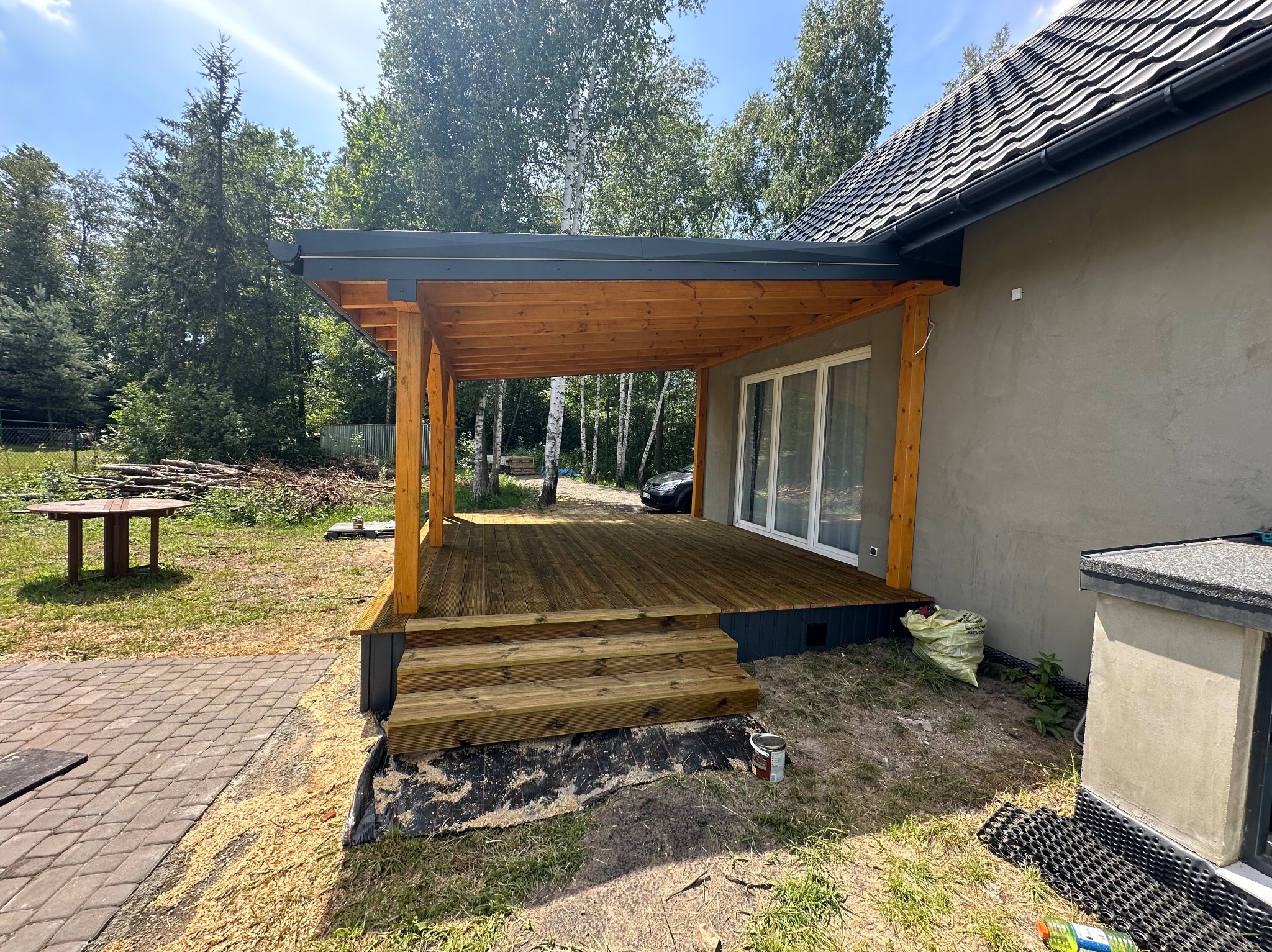 Zadaszony taras z drewnianą podłogą i schodami, przylegający do budynku z szarą elewacją. Widoczne drzewa w tle i elementy wykończeniowe.