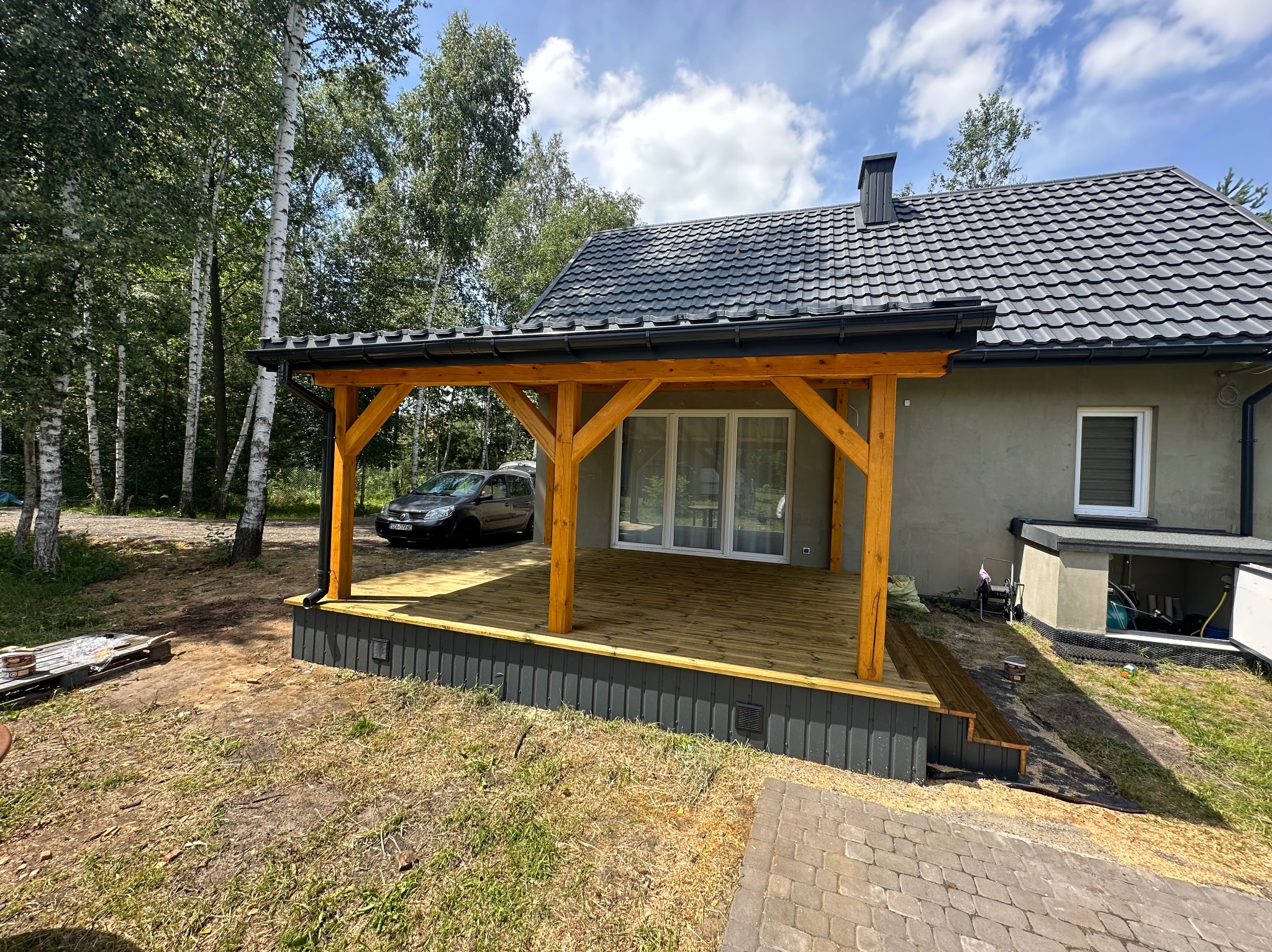 Drewniany taras z zadaszeniem przy domu z szarą elewacją i grafitowym dachem. Widoczne drzewa i samochód w tle. Nowoczesna architektura.