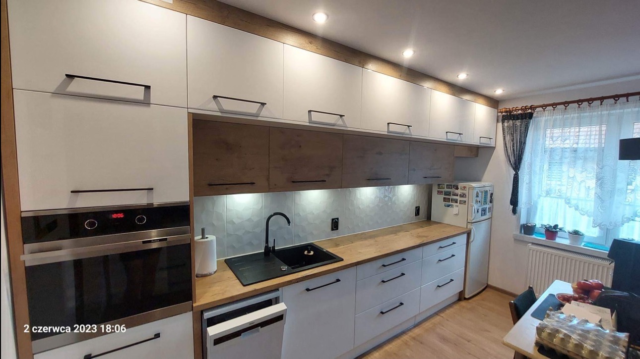 Nowoczesna kuchnia z białymi i drewnianymi szafkami, czarnym zlewem i armaturą. Uwagę przykuwają heksagonalne płytki nad blatem i oświetlenie LED.