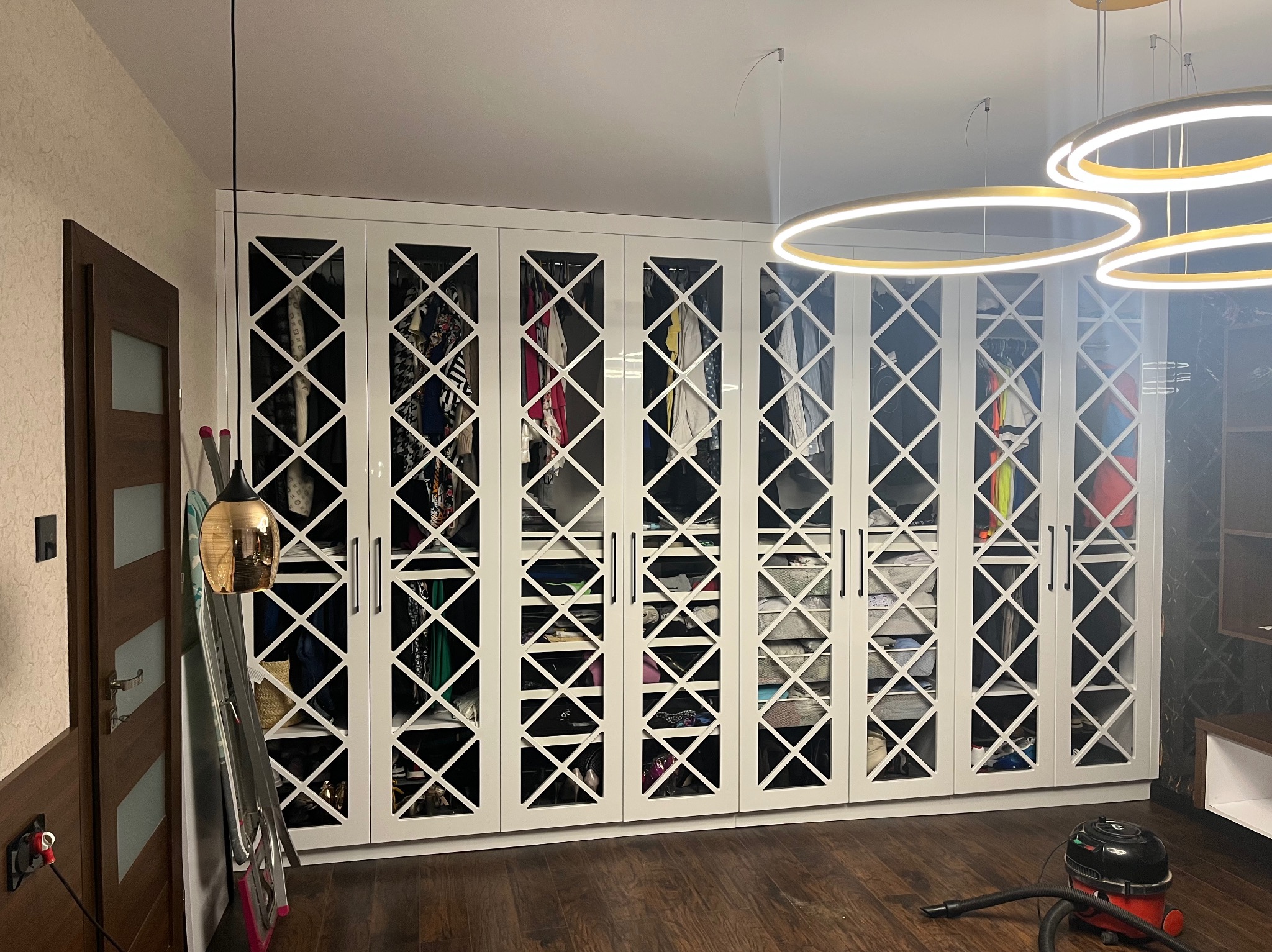Biała, zabudowana szafa z dekoracyjnymi, ażurowymi drzwiami w nowoczesnym wnętrzu. Widoczne ubrania i akcesoria wewnątrz. Na podłodze odkurzacz.