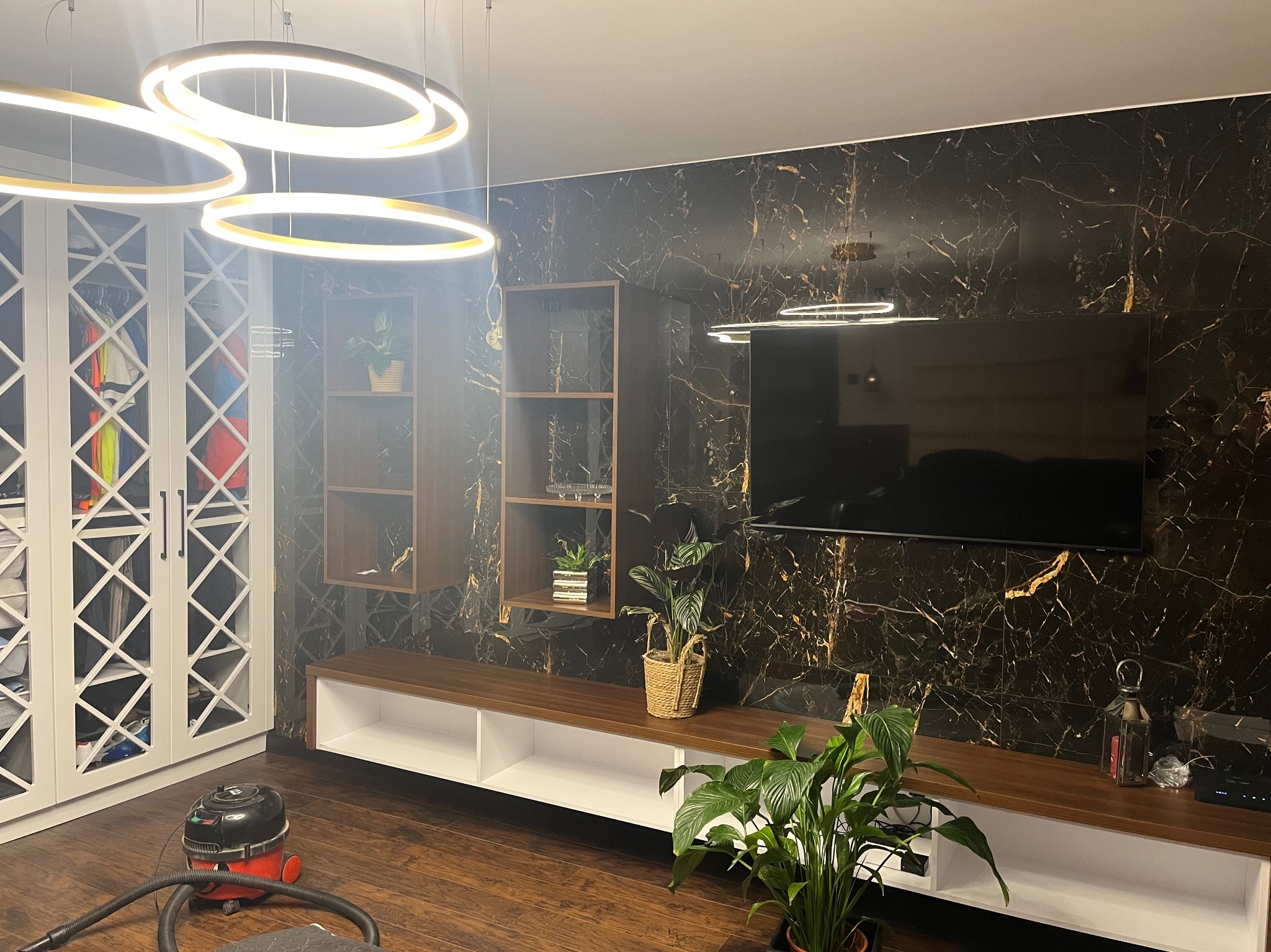 Eleganckie wnętrze: ściana z czarnego marmuru z telewizorem, biała szafa z dekoracyjnymi wstawkami, drewniana podłoga i nowoczesne oświetlenie.