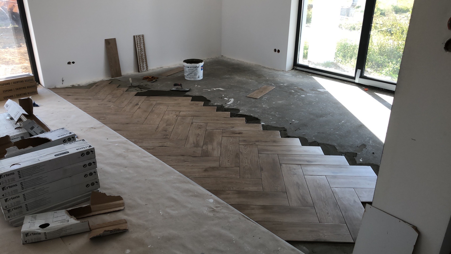 Układanie płytek podłogowych w jodełkę. Częściowo ułożona podłoga z płytek imitujących drewno, widoczne paczki płytek i narzędzia glazurnicze. Jasne wnętrze z dużym oknem.