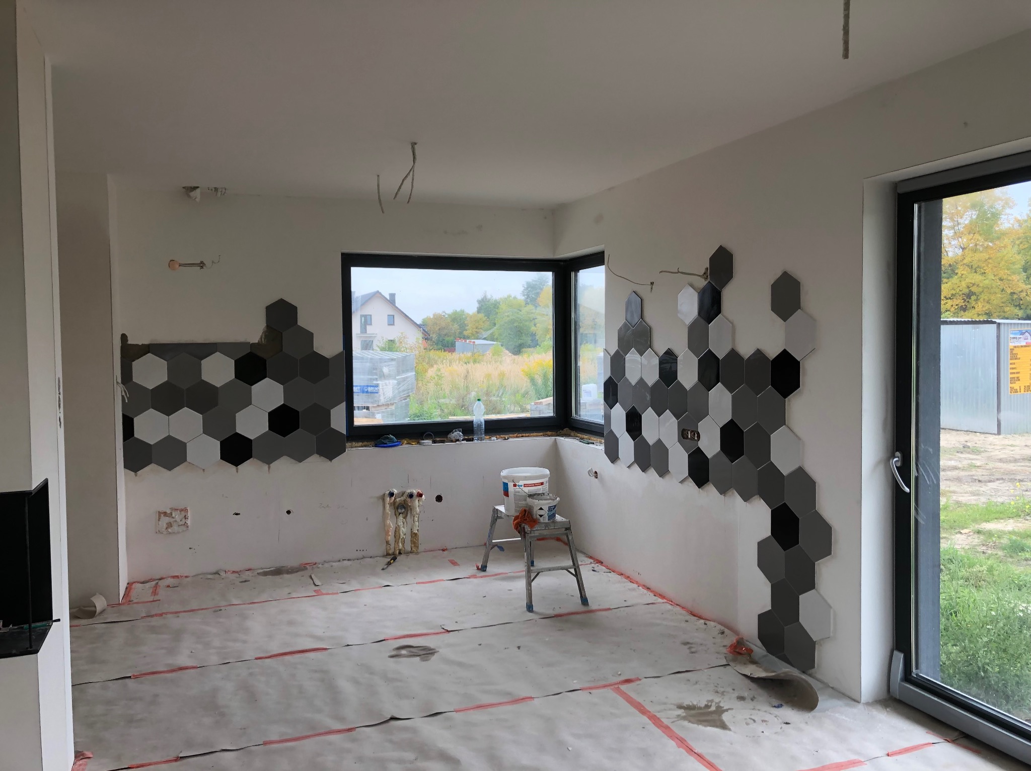 Wnętrze w trakcie układania heksagonalnych płytek w odcieniach szarości i czerni na ścianie. Widoczne narzędzia i materiały, okno z widokiem na zewnątrz.