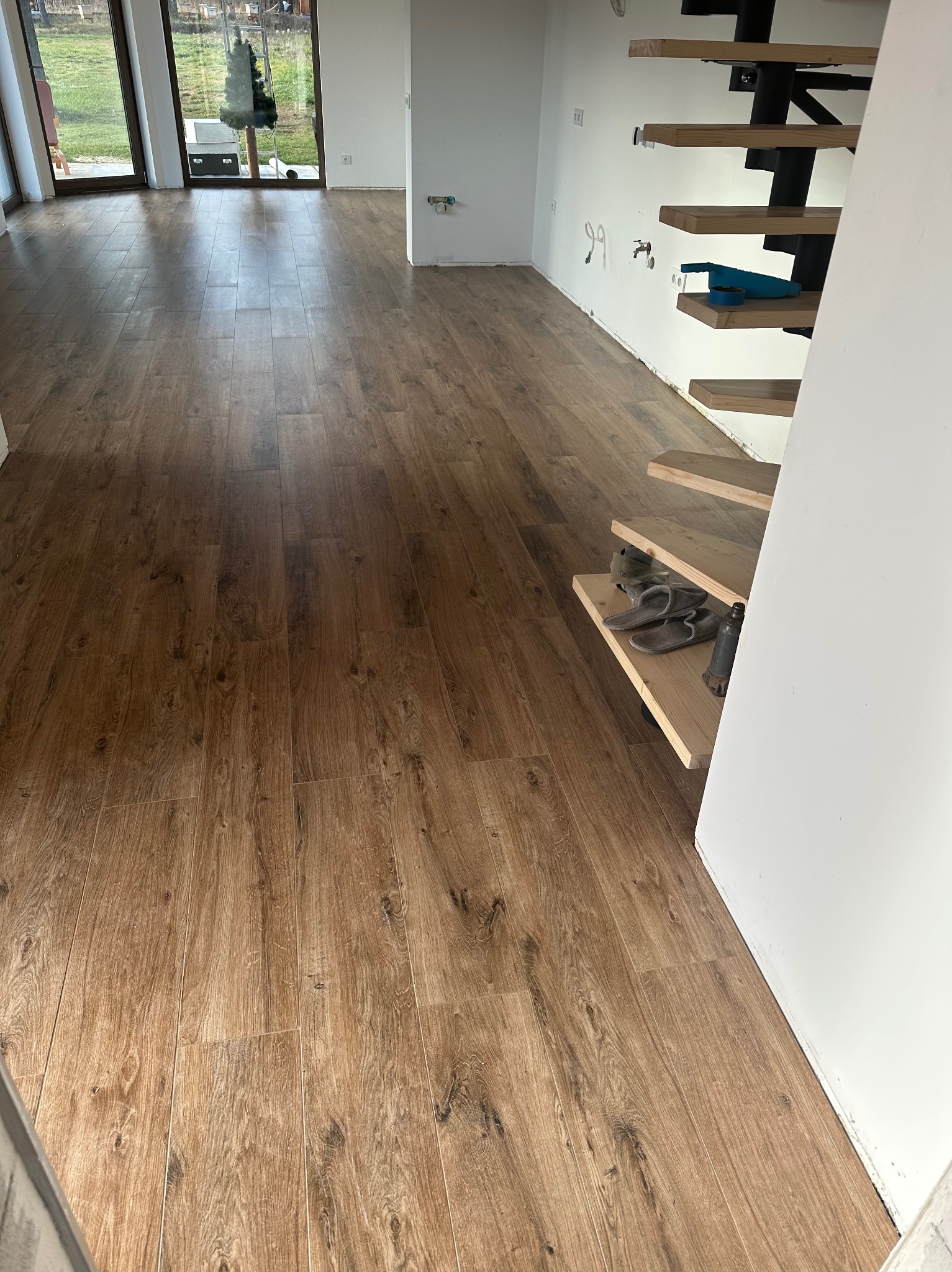 Ułożone panele podłogowe w odcieniach brązu, imitujące drewno, w jasnym wnętrzu z widocznymi schodami i dużymi oknami. Widok na ogród przez okno.