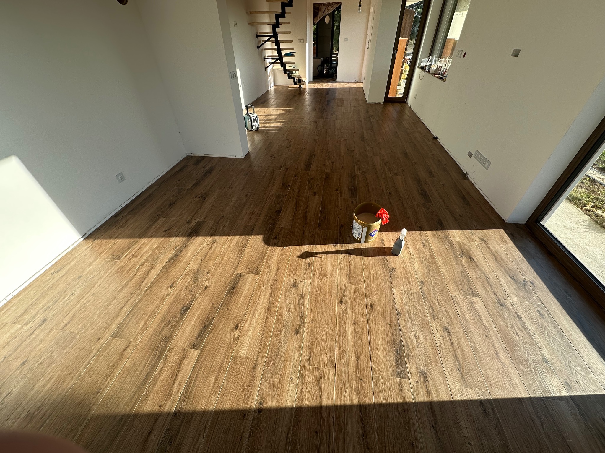 Ukończona podłoga z paneli imitujących drewno w jasnym, przestronnym wnętrzu z białymi ścianami i schodami. Widoczne narzędzia i wiadro po środkach do pielęgnacji.