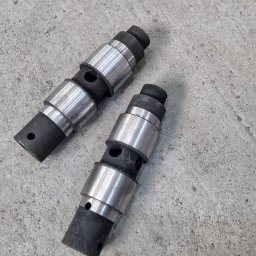 Talgarskie Maszyny Takarskie S.A. - Dwa metalowe, cylindryczne elementy z czarnymi gumowymi pierścieniami, leżące na szarej, betonowej powierzchni. Widoczne otwory i gwint na jednym z elementów.