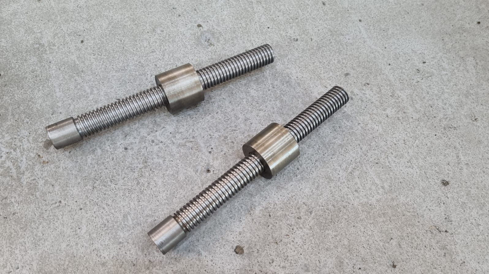 Dwa precyzyjnie wykonane, metalowe wały śrubowe z nakrętkami, leżące na szarej, betonowej powierzchni. Widoczne ślady obróbki skrawaniem.
