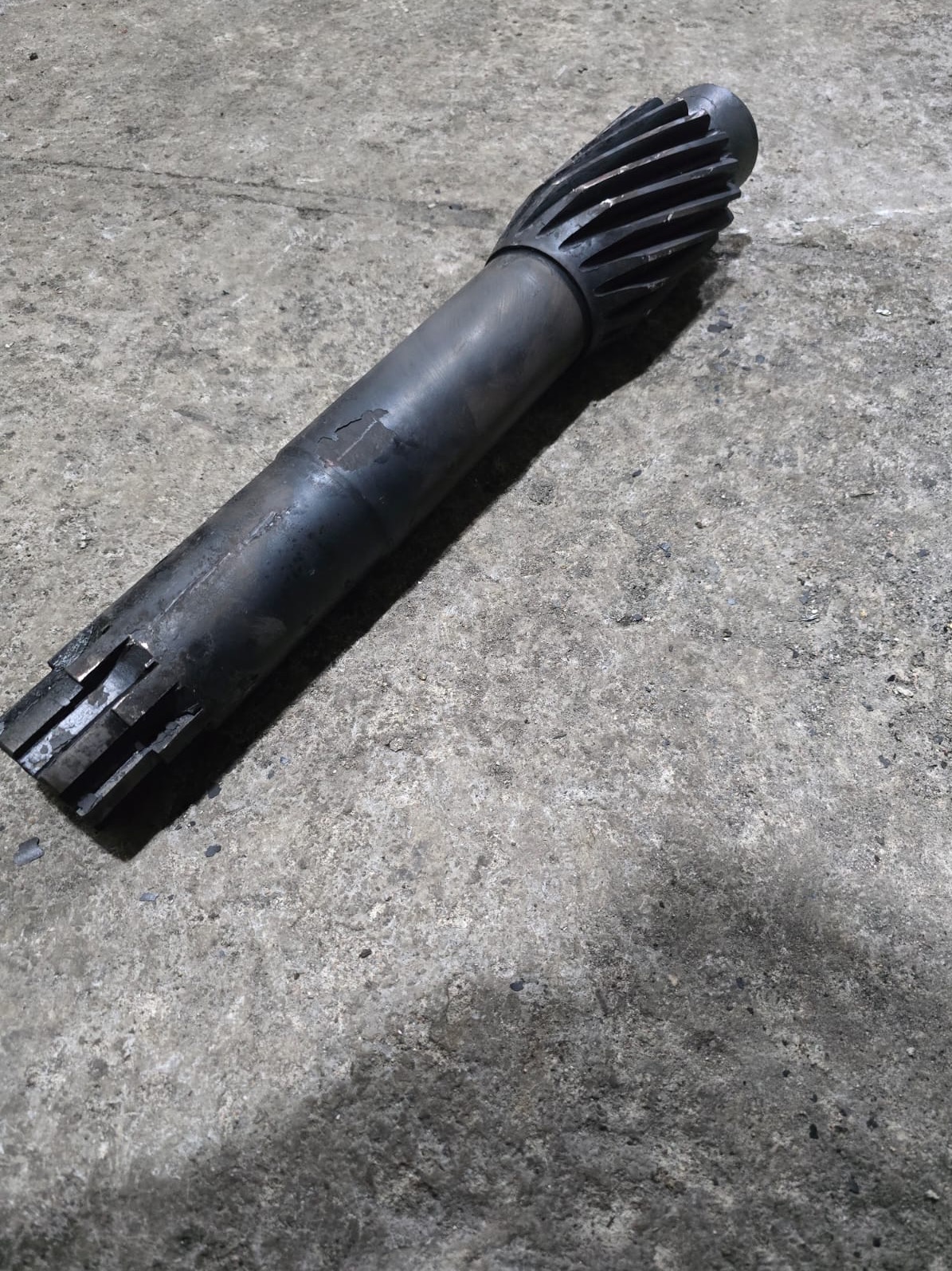 Metalowy wał z zębami i rowkami, leżący na szarym, betonowym podłożu. Widoczne ślady obróbki i niewielkie uszkodzenia powierzchni.