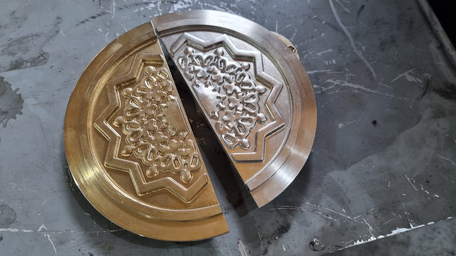 Dwie połówki okrągłego, metalowego elementu z wyfrezowanym wzorem kwiatowym i gwiazdą, leżące na szarej, roboczej powierzchni. Widoczne ślady obróbki.