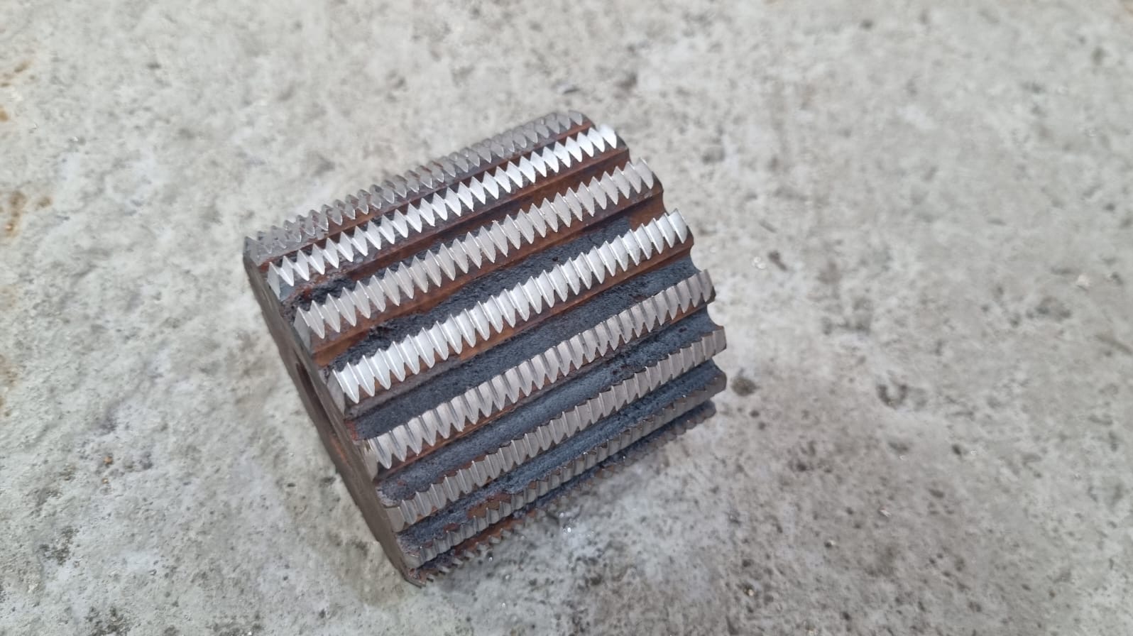 Metalowy frez ślimakowy z wyraźnymi zębami, leżący na szarej, betonowej powierzchni. Widoczne ślady użytkowania i drobne zabrudzenia.