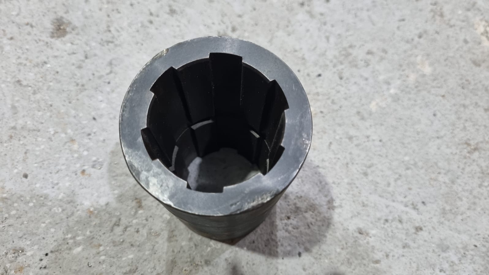 Metalowy element cylindryczny z wewnętrznymi wycięciami, leżący na szarej, betonowej powierzchni. Widoczne ślady obróbki i delikatne niedoskonałości materiału.