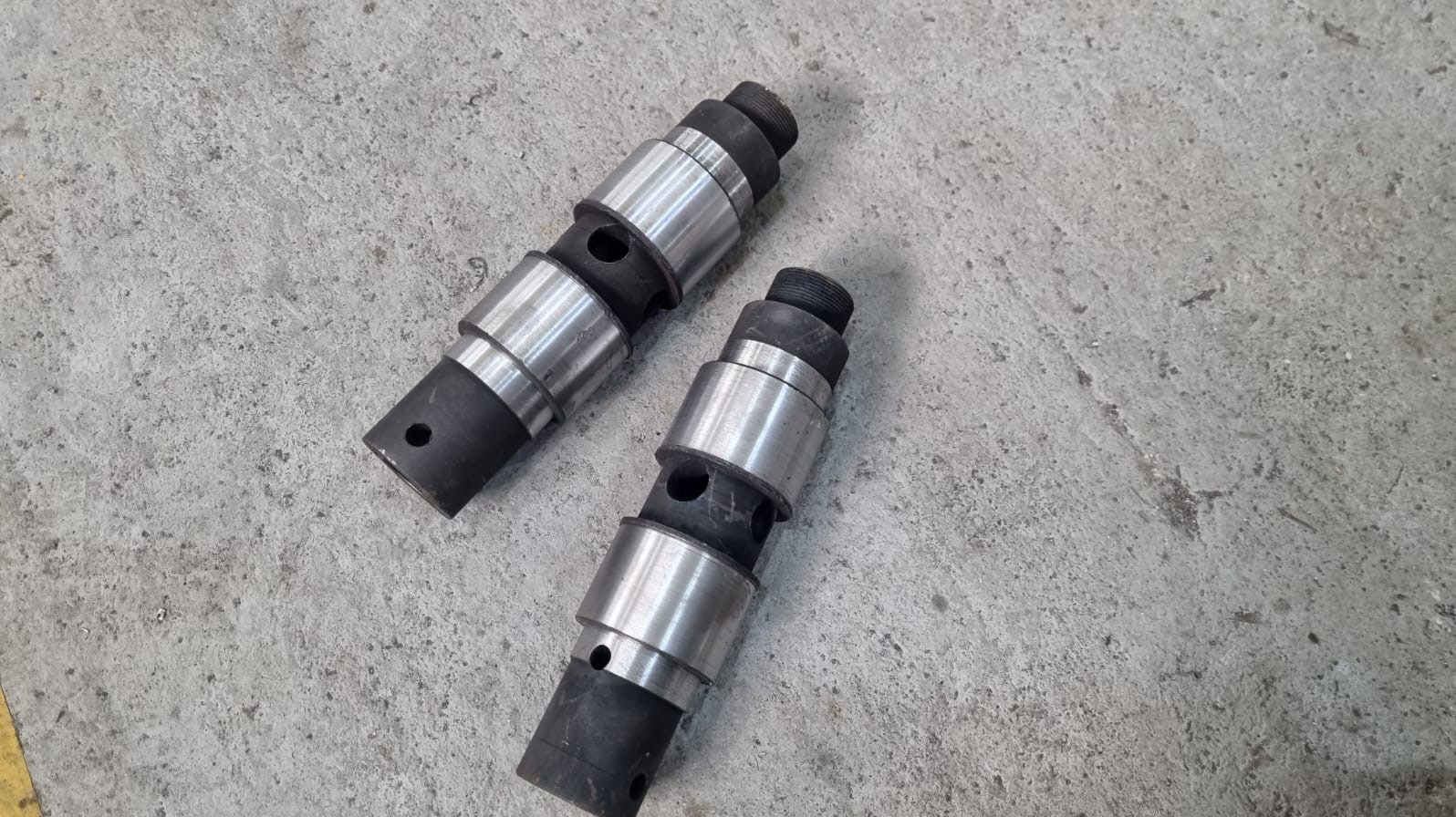 Dwa metalowe, cylindryczne elementy z czarnymi gumowymi pierścieniami, leżące na szarej, betonowej powierzchni. Widoczne otwory i gwint na jednym z elementów.