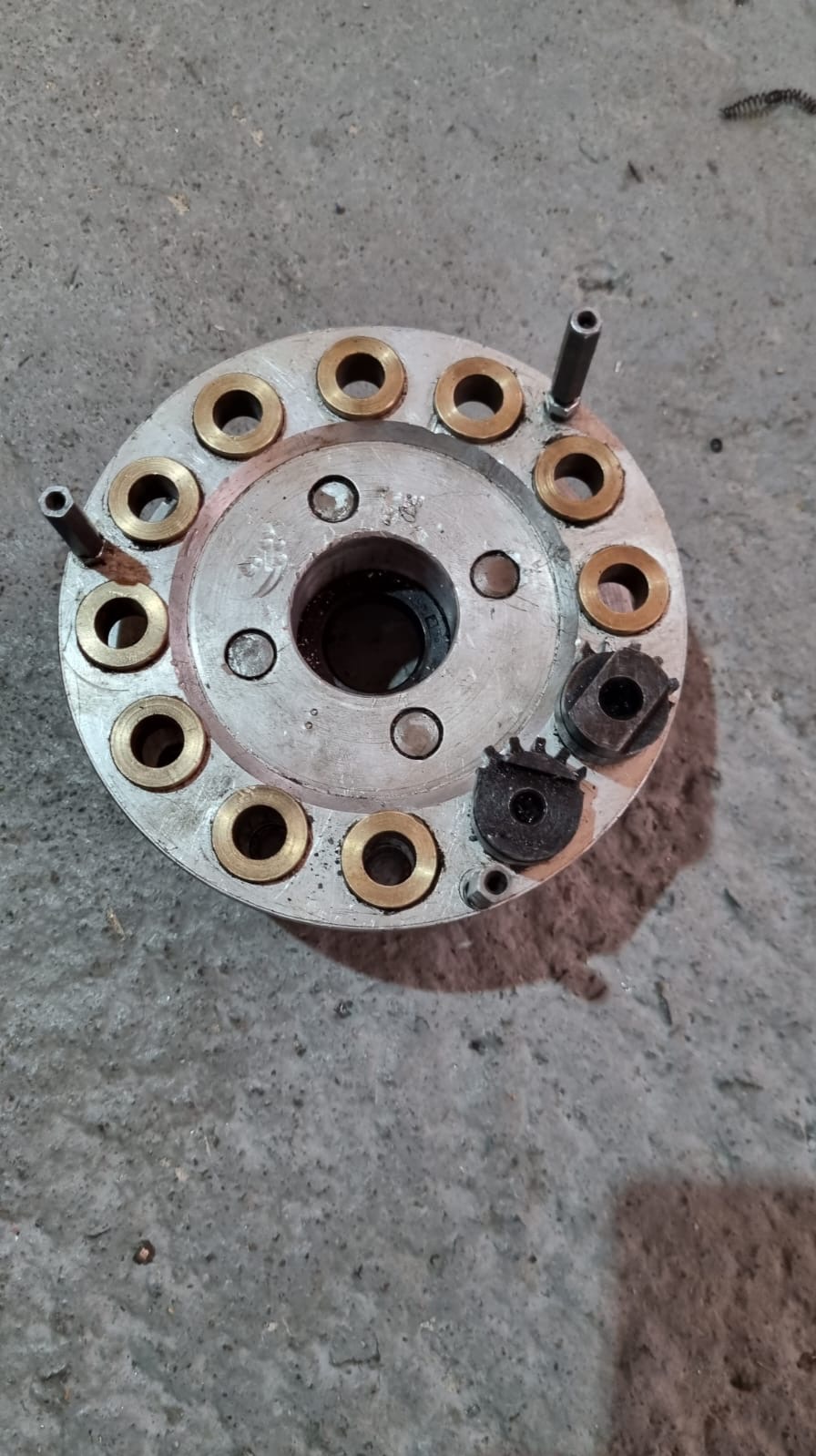 Szara, okrągła metalowa tarcza z mosiężnymi tulejami i czarnymi zębatkami na betonowym tle. Widoczne ślady obróbki i zużycia.