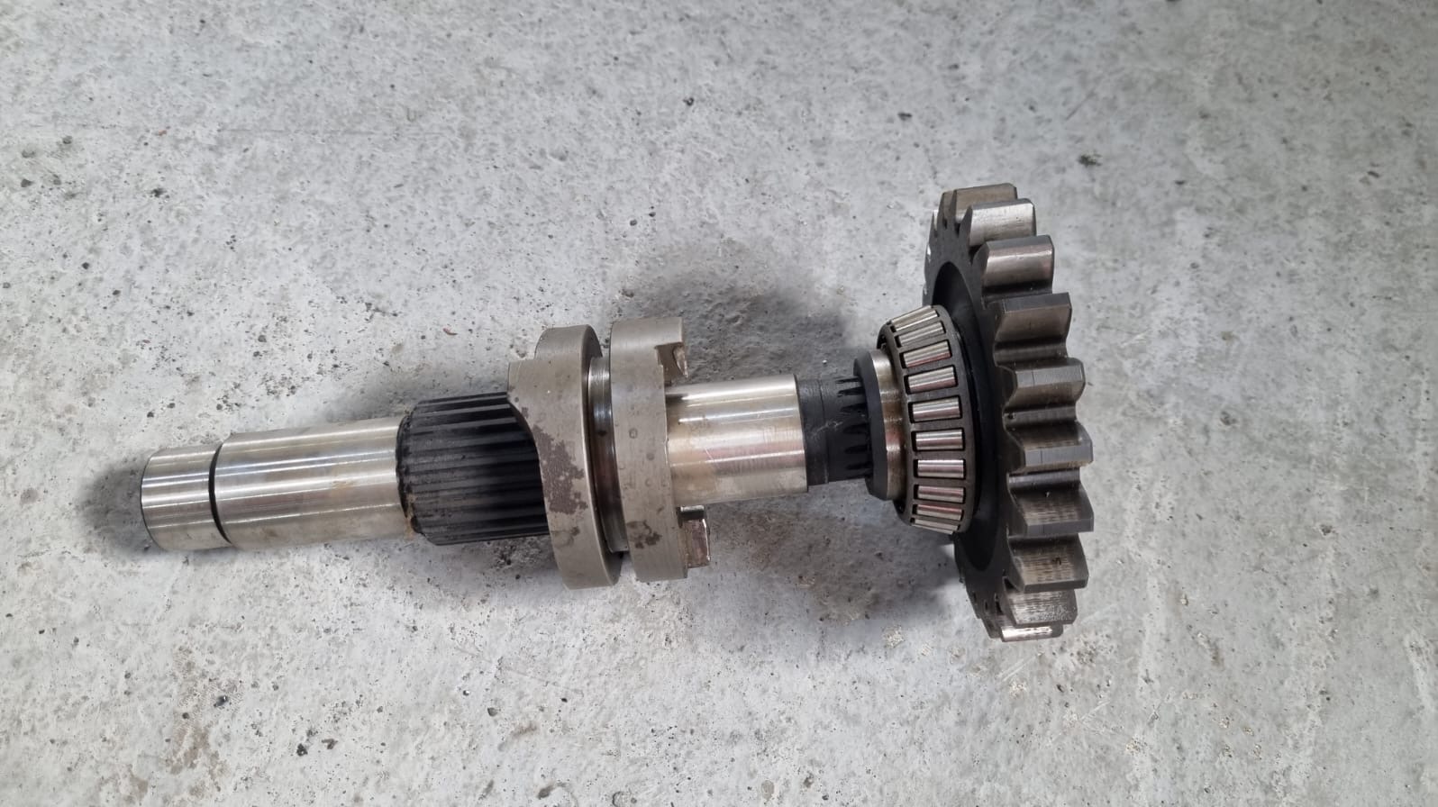 Metalowy wał z zębatką i łożyskiem stożkowym na szarym tle. Widoczne ślady obróbki i montażu. Element mechaniczny po procesie toczenia i frezowania.