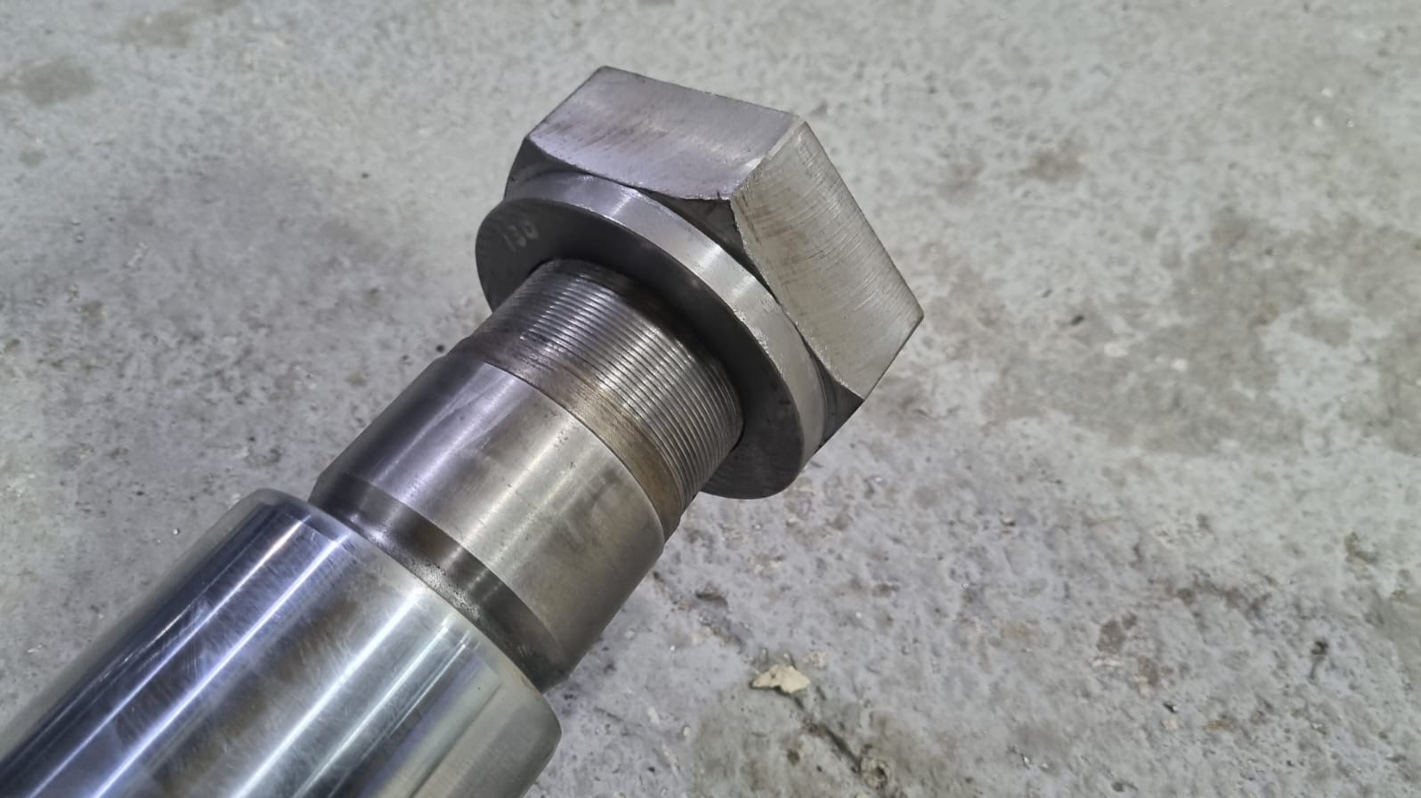 Metalowy wał z gwintem i nakrętką sześciokątną, leżący na szarej, betonowej powierzchni. Widoczne ślady obróbki i delikatne refleksy światła na metalu.