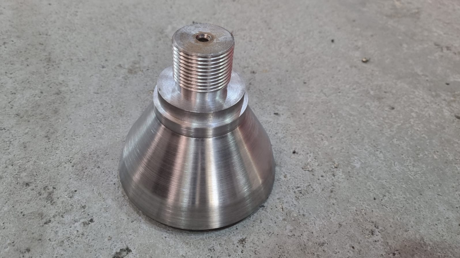 Metalowy element z gwintem i otworem, wykonany na tokarce. Widoczne ślady obróbki skrawaniem. Detal na szarym, betonowym tle. Precyzyjna obróbka metalu.