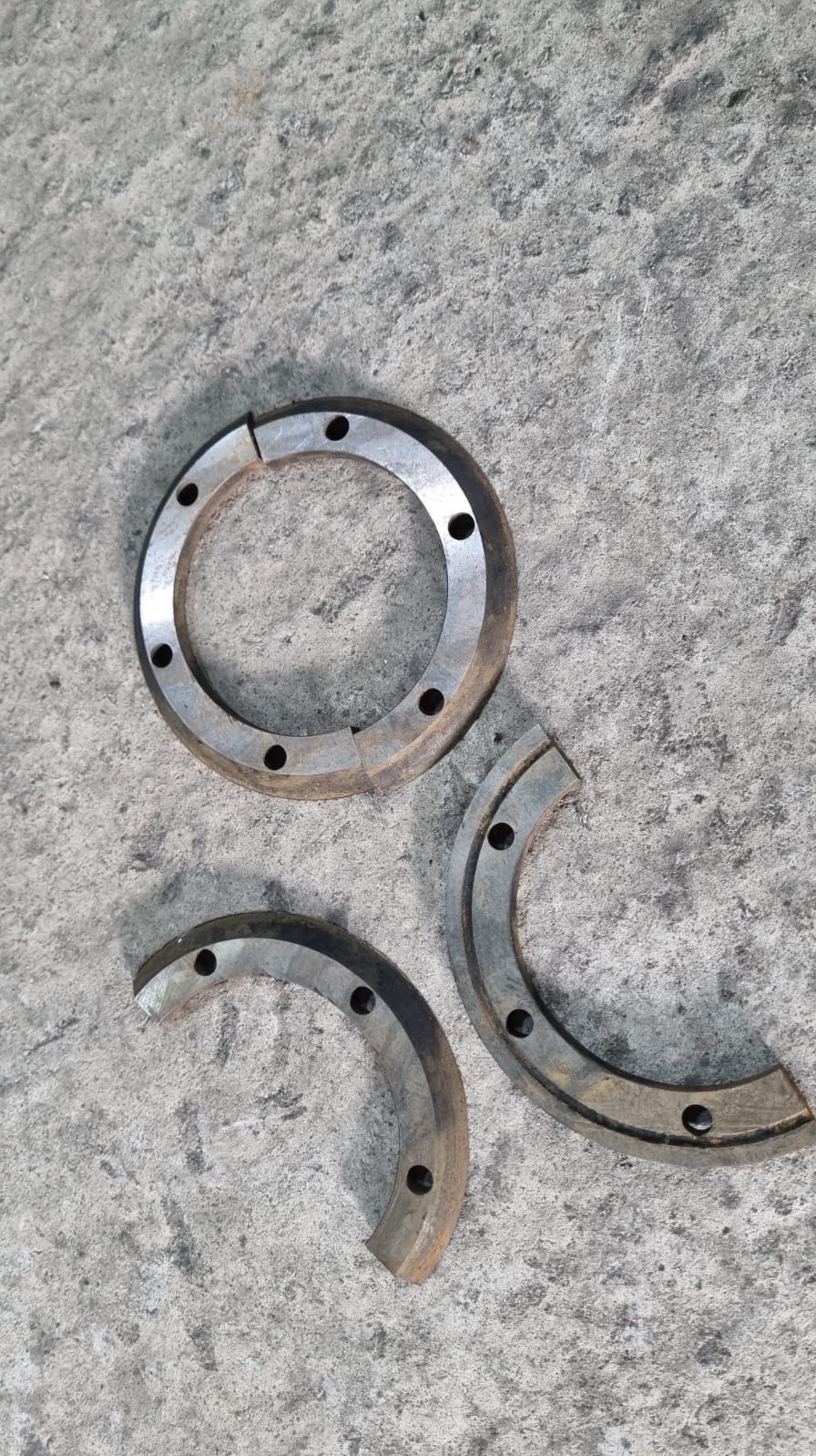 Metalowe pierścienie z otworami na szarym, betonowym tle. Jeden pierścień jest kompletny, dwa pozostałe to jego fragmenty. Widoczne ślady obróbki.