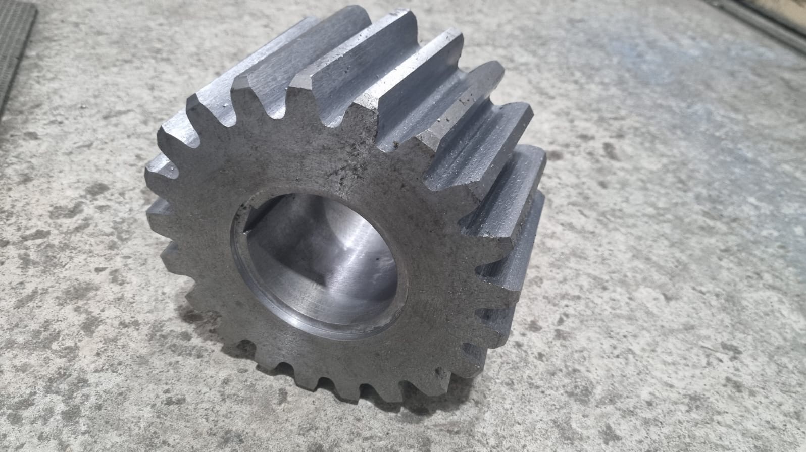 Metalowe koło zębate z otworem w środku, leżące na szarej, betonowej powierzchni. Widoczne ślady obróbki.