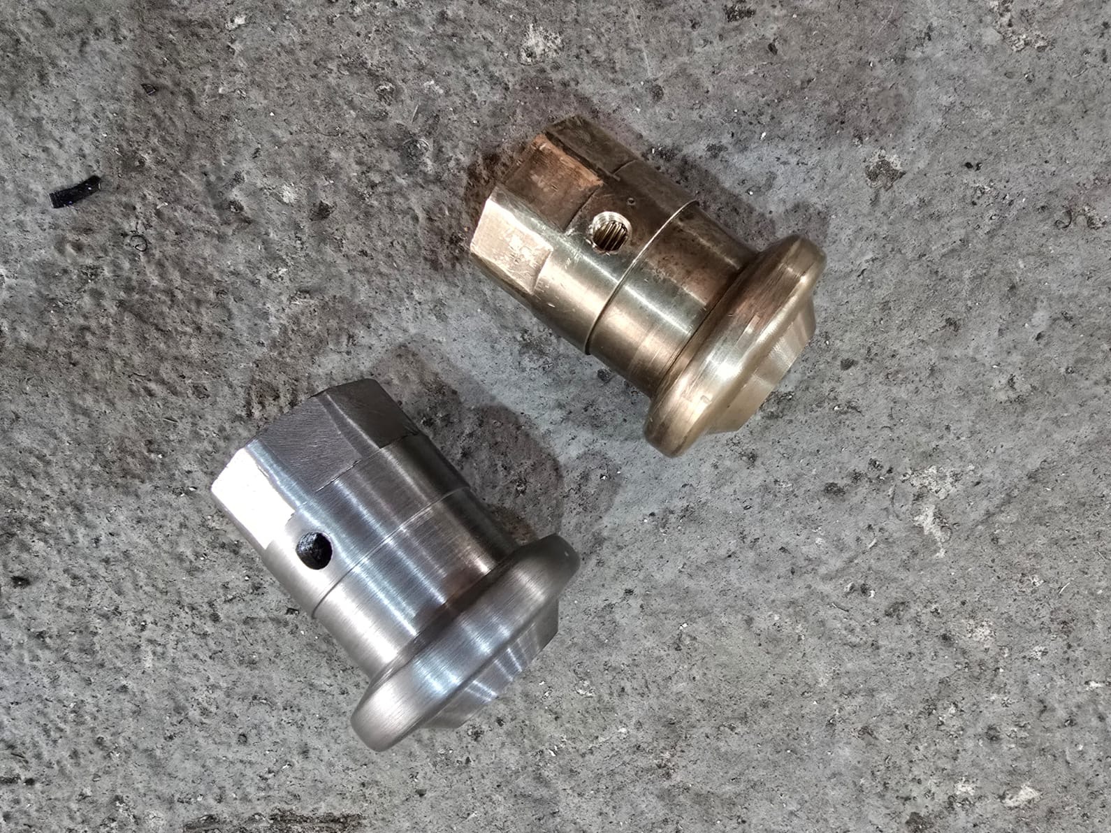 Dwa metalowe elementy cylindryczne z kołnierzami, jeden srebrny, drugi mosiężny, leżą na szarej, betonowej powierzchni. Widoczne otwory i ślady obróbki.