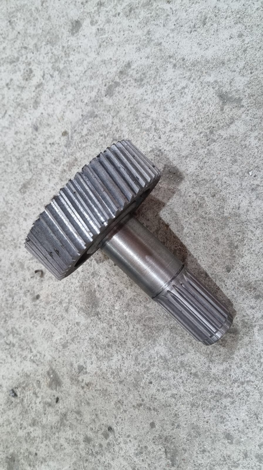 Metalowa zębatka z wałem na szarym, betonowym tle. Widoczne ślady obróbki. Detal z bliska, ukazujący precyzję wykonania i solidność materiału.