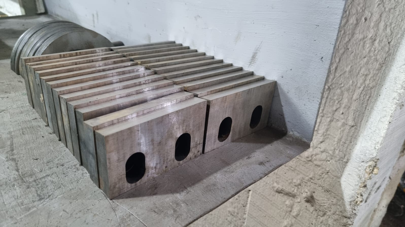 Rząd metalowych bloków z otworami, ułożonych na betonowej powierzchni. Widoczne ślady obróbki, szlifowania. W tle okrągłe, metalowe elementy.