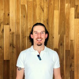 Zmasowane Dobro - powięziowy masaż Gdańsk - Uśmiechnięty mężczyzna z brodą i długimi włosami w białej koszulce polo na tle drewnianej ściany. Widoczny tatuaż na ramieniu i mikrofon przypięty do koszulki.