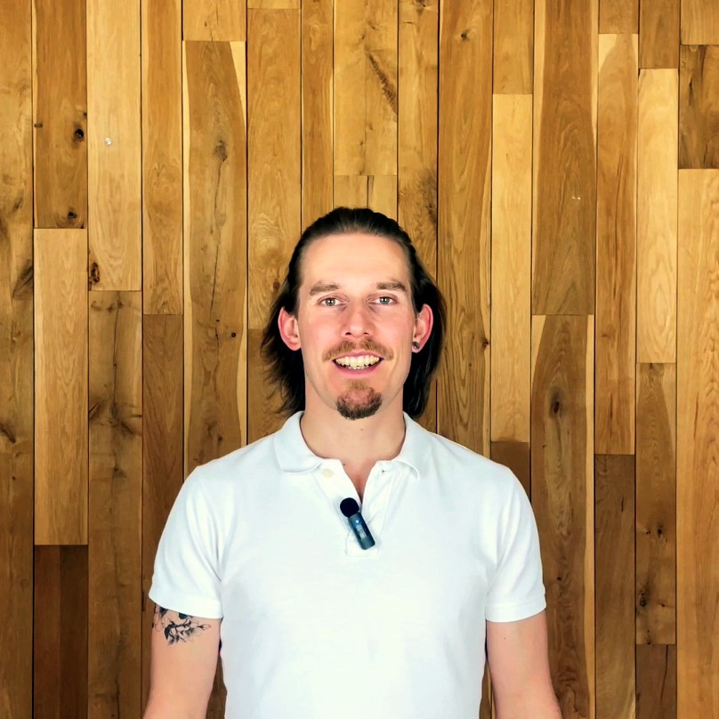 Uśmiechnięty mężczyzna z brodą i długimi włosami w białej koszulce polo na tle drewnianej ściany. Widoczny tatuaż na ramieniu i mikrofon przypięty do koszulki.