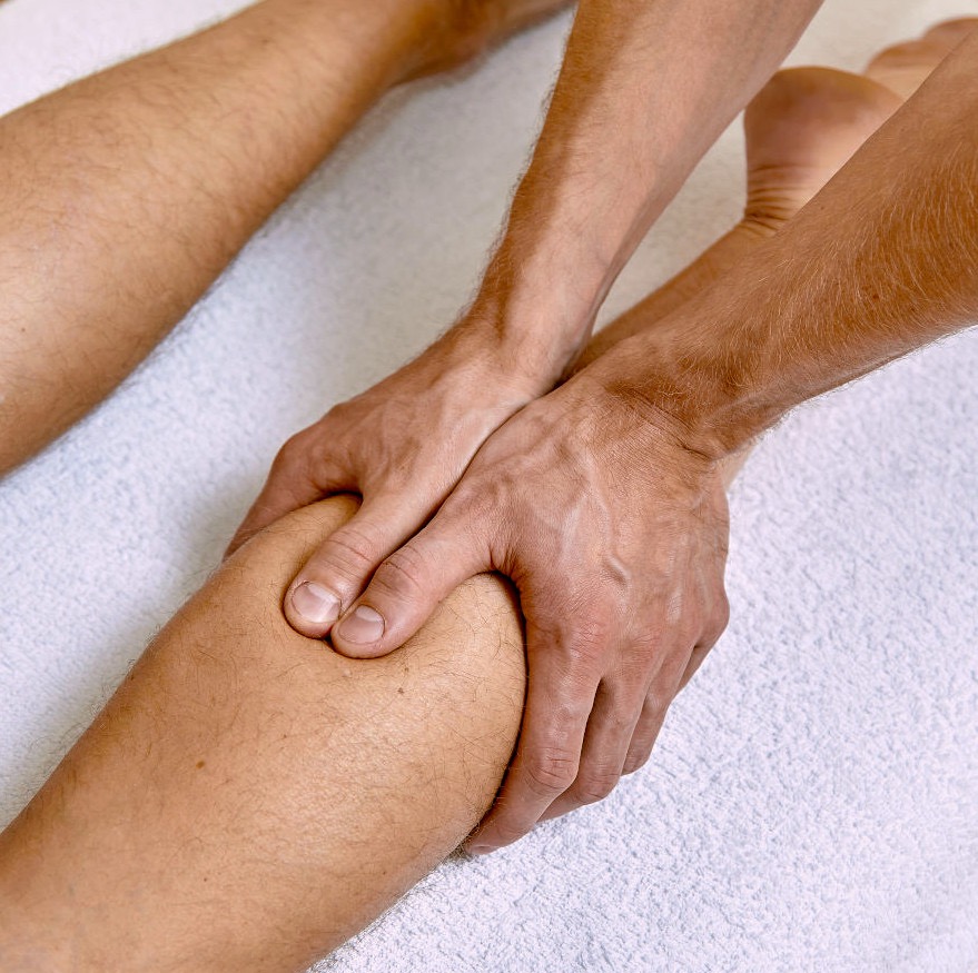 Dłonie terapeuty uciskają łydkę pacjenta na białym ręczniku. Bliski kadr, skupienie na manualnej terapii mięśni. Delikatne światło podkreśla teksturę skóry.