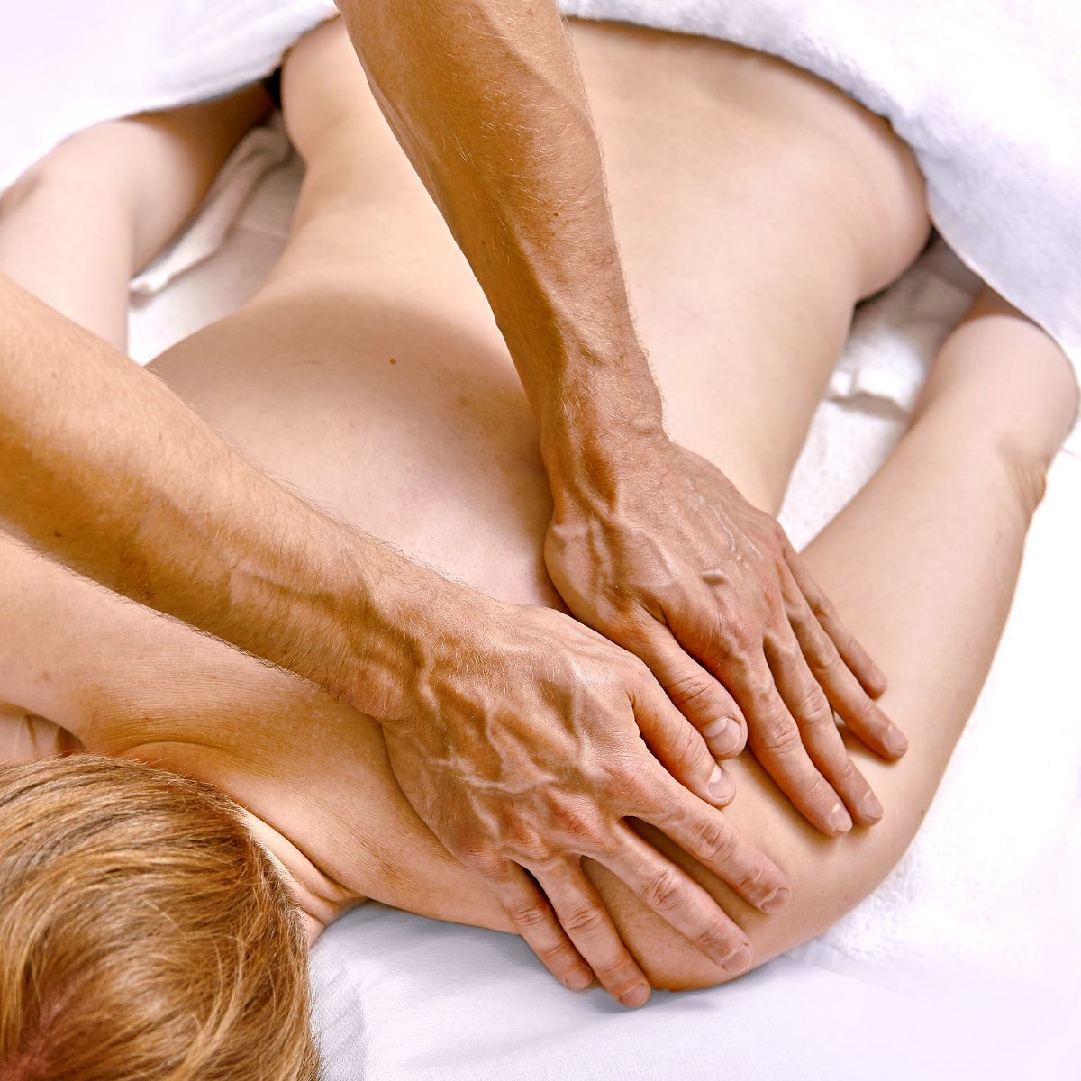 Dłonie masujące plecy kobiety leżącej na białym ręczniku. Widoczne detale skóry i napięcie mięśni. Jasne, naturalne oświetlenie podkreśla anatomię.