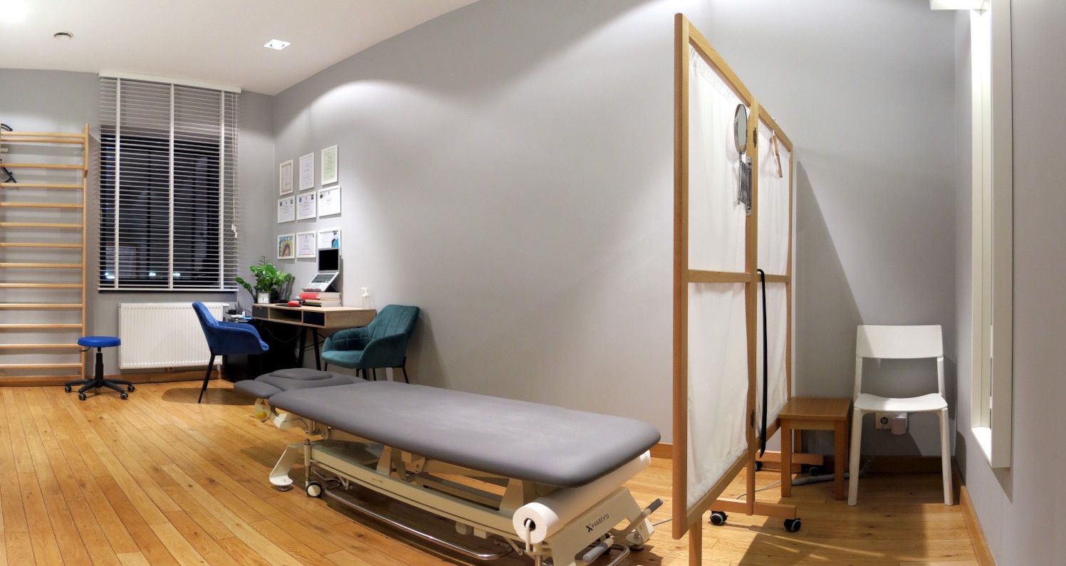 Jasne wnętrze gabinetu z drewnianą podłogą. Centralnie stół do zabiegów, w tle biurko z laptopem, drabinka gimnastyczna i parawan. Stonowana kolorystyka.