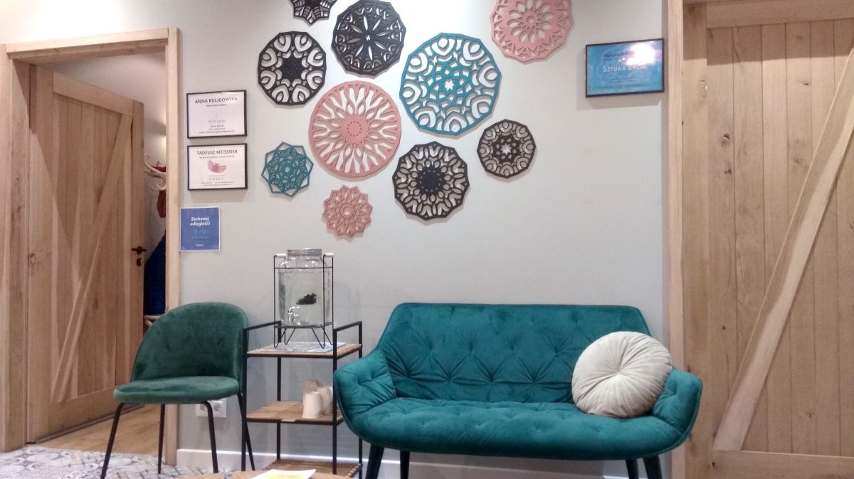 Przytulne wnętrze gabinetu z turkusową sofą i dekoracjami na ścianie. Widoczne certyfikaty i akwarium z rybką. Drewniane drzwi dodają ciepła.