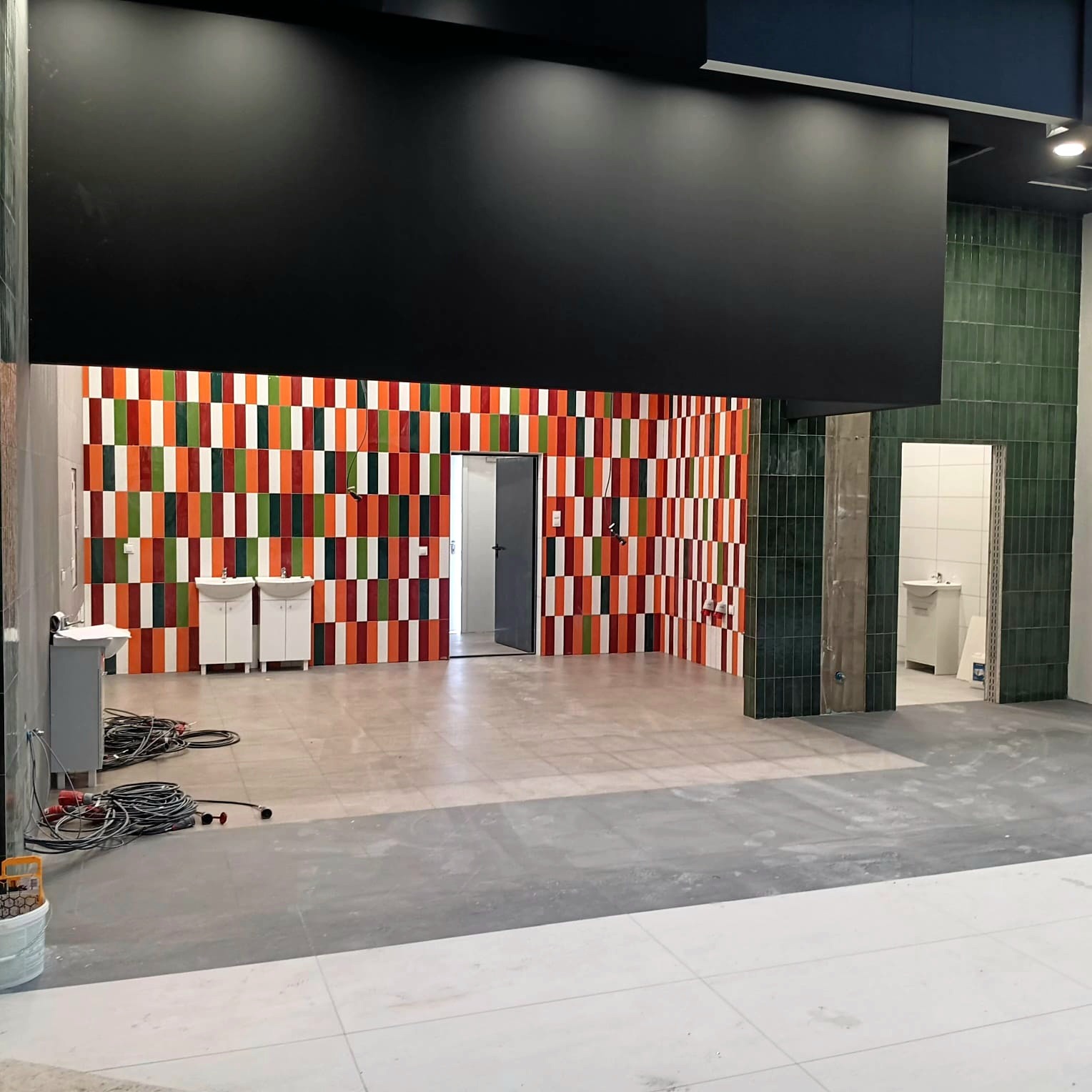 Wnętrze komercyjne w trakcie wykańczania: kolorowa mozaika z płytek na ścianie, surowe podłogi, umywalki i narzędzia. Widok na sąsiednie pomieszczenie z zielonymi płytkami.