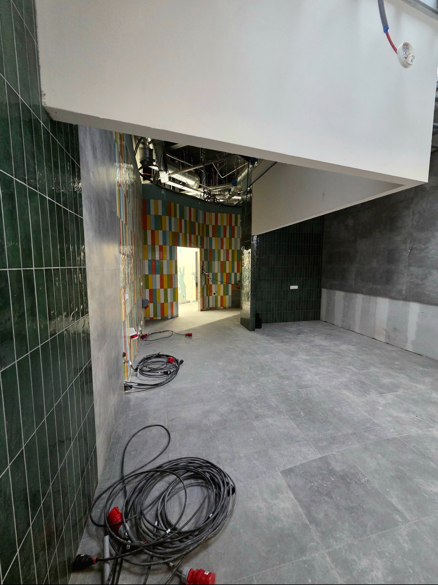 Wnętrze w trakcie wykańczania: szare płytki podłogowe, ściany z zielonej i kolorowej glazury, widoczne kable i instalacje. Surowy stan, przygotowanie do dalszych prac.