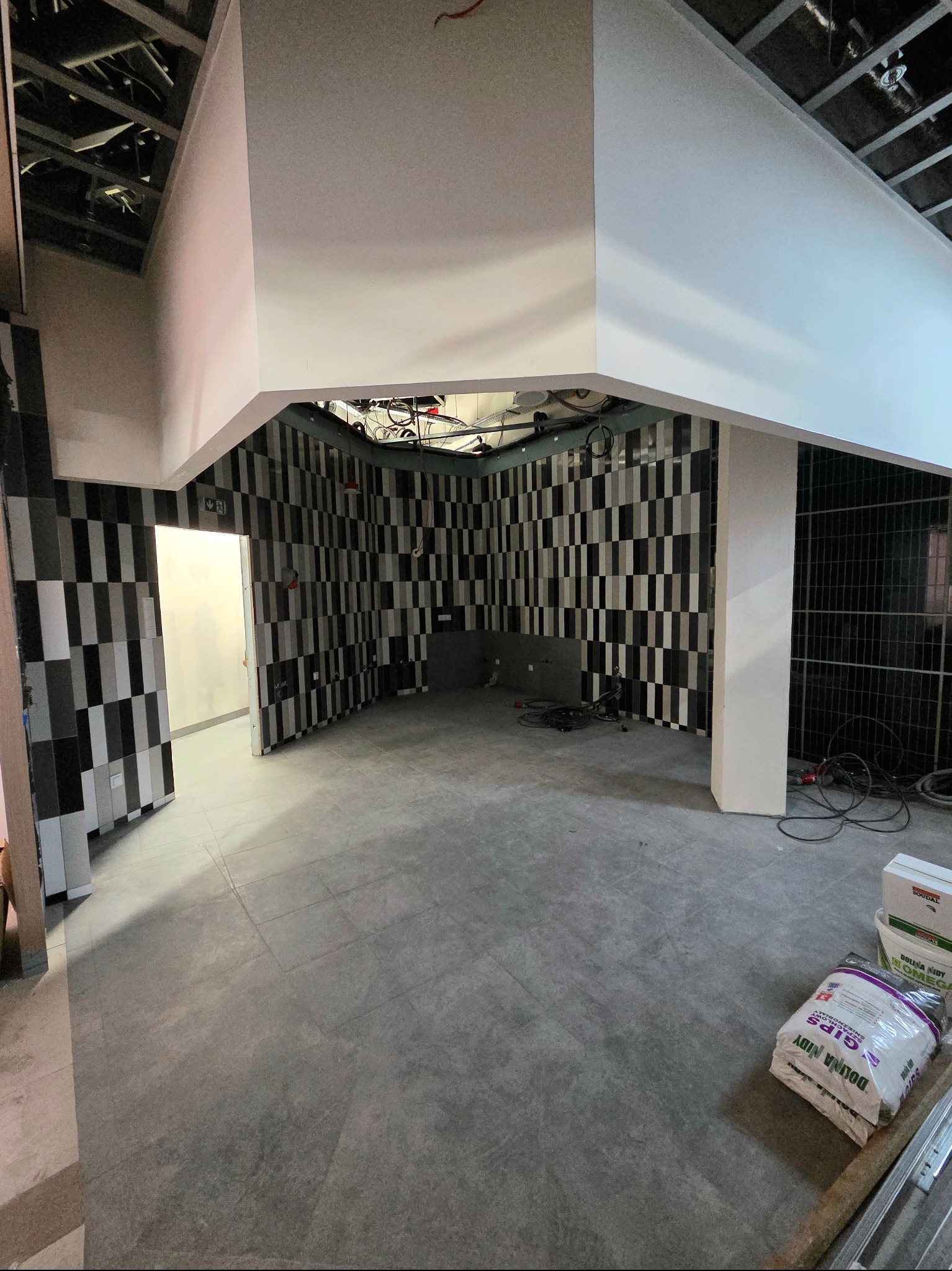 Wnętrze w trakcie wykańczania: szara podłoga z płytek, ściany z czarno-białą szachownicą, widoczne kable i materiały budowlane. Surowy, nowoczesny design.