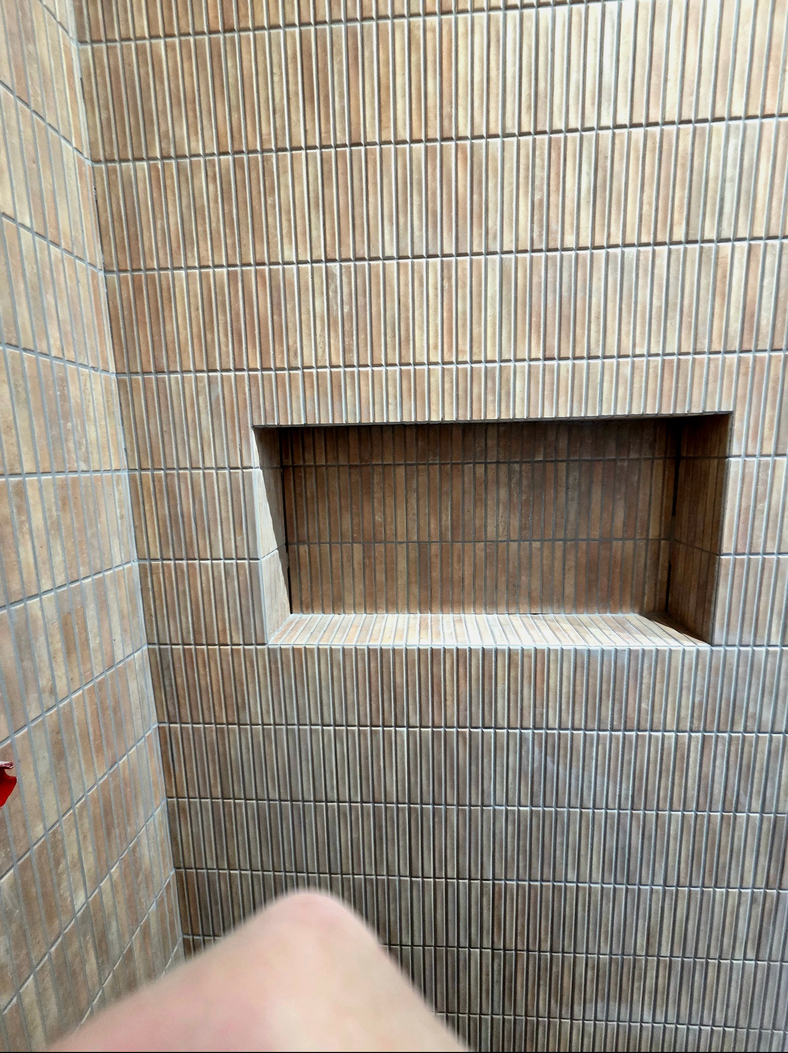 Beżowa ściana w łazience wyłożona wąskimi, pionowymi płytkami z wnęką. Widoczny fragment ciała w dolnej części kadru.