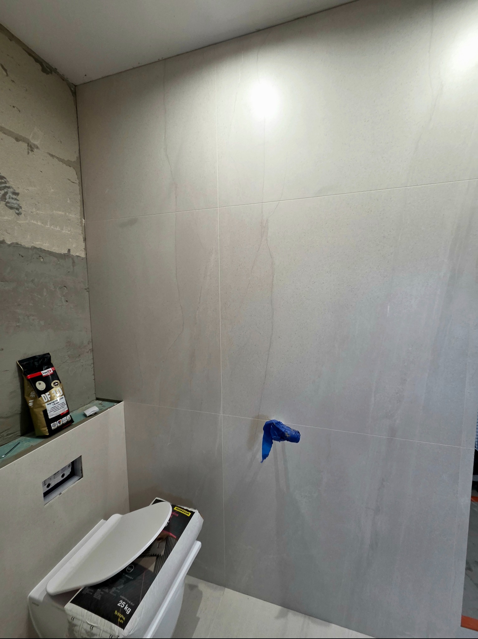 Jasne, matowe płytki na ścianie łazienki, częściowo wykończone. Widoczny stelaż podtynkowy WC, worek z klejem i niebieska folia zabezpieczająca.