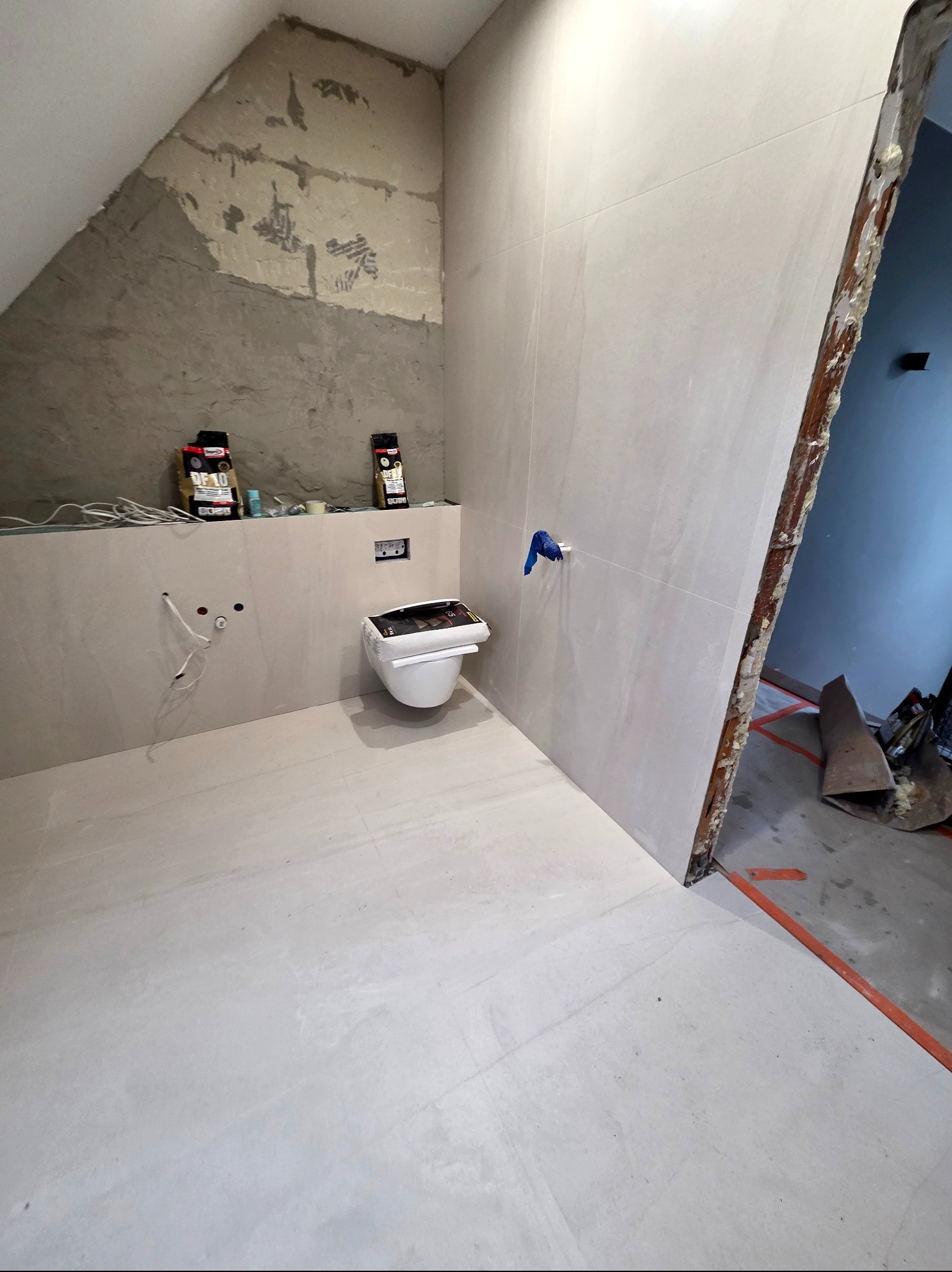 Jasna łazienka w trakcie wykańczania: białe płytki na ścianach i podłodze, podwieszana toaleta, surowy fragment ściany pod skosem. Widoczne materiały budowlane.