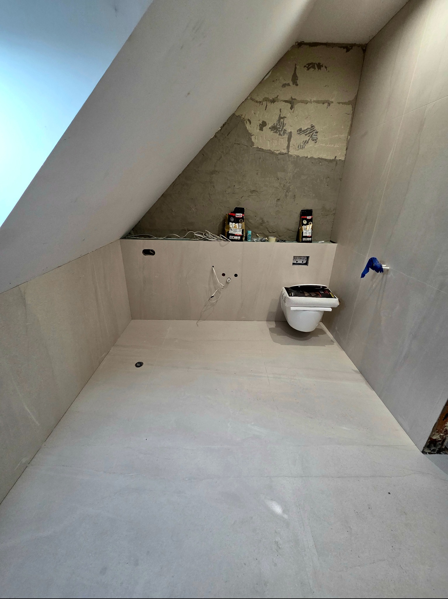 Łazienka na poddaszu w trakcie wykańczania: jasne płytki, częściowo wykończona ściana, podwieszana toaleta i materiały budowlane. Widok na wnętrze w trakcie prac.