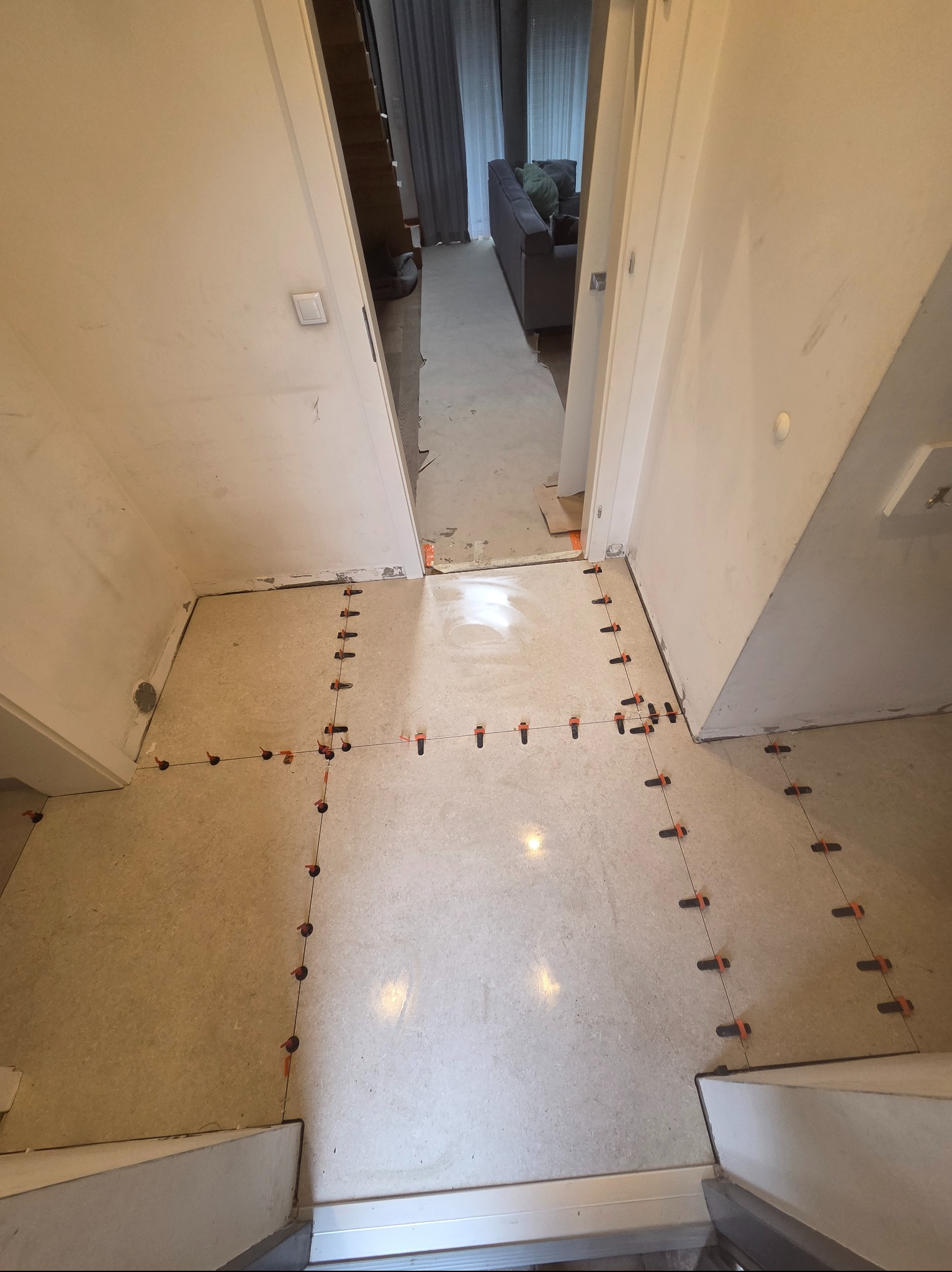 Układanie jasnych płytek podłogowych z użyciem systemu poziomowania. Widoczne krzyżyki dystansowe i kliny. W tle widoczny fragment salonu z szarą sofą.