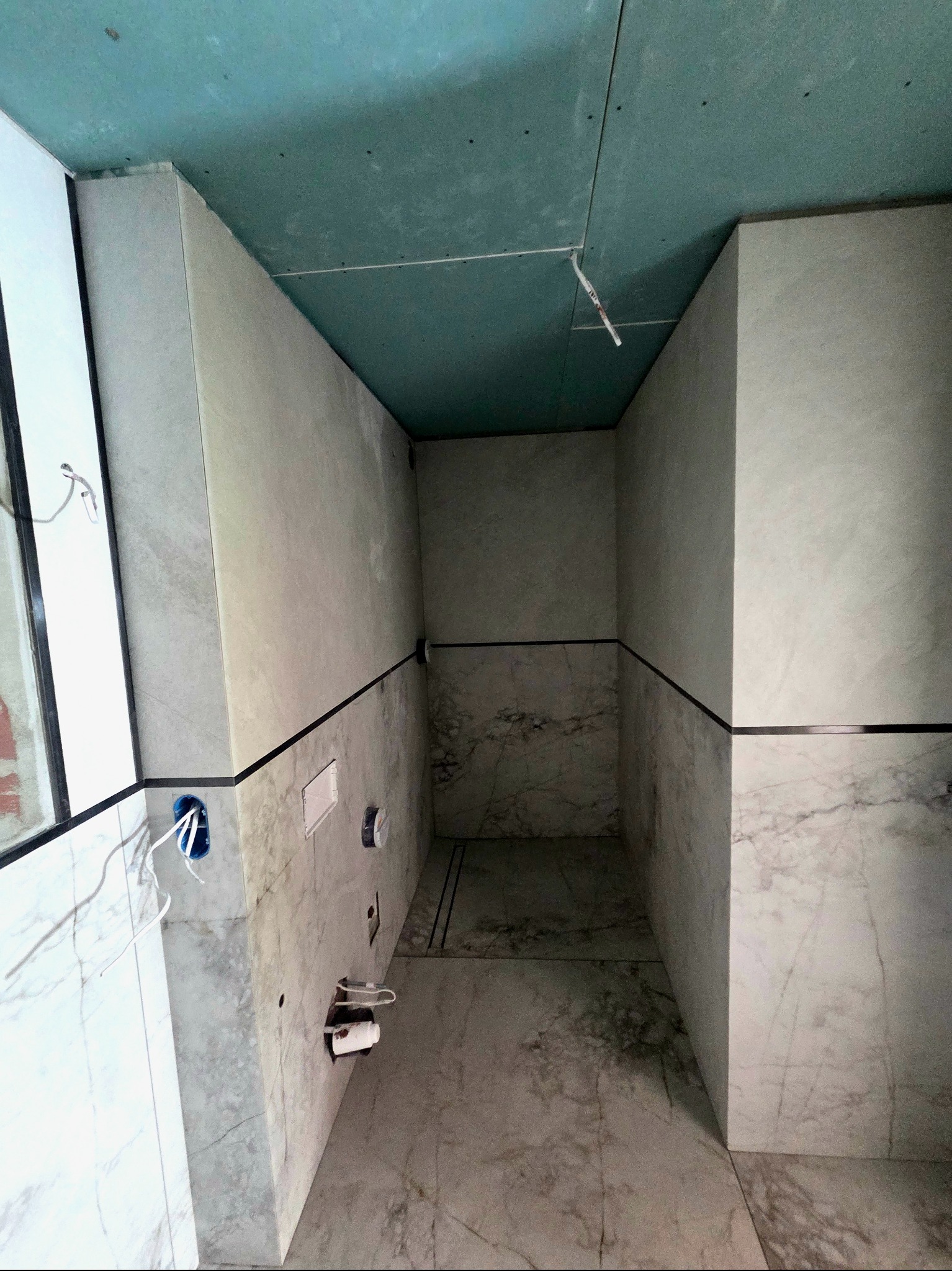Wnętrze łazienki w trakcie wykańczania: marmurowe płytki na ścianach i podłodze, sufit z płyt gipsowo-kartonowych, widoczne instalacje elektryczne i sanitarne.