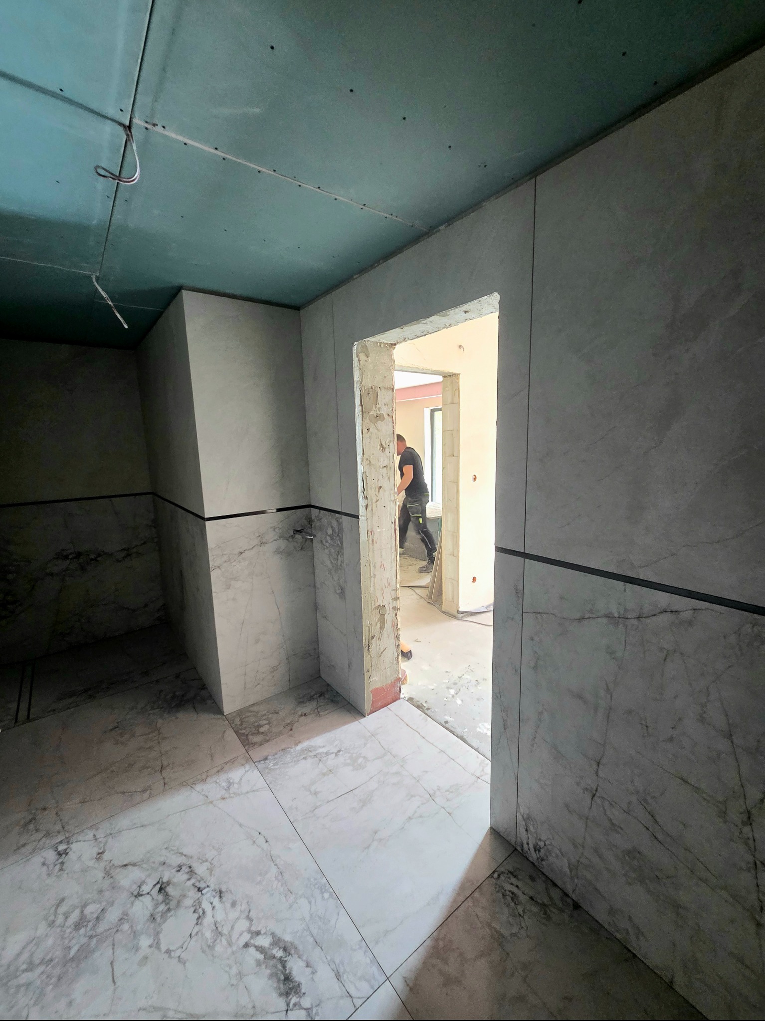 Wnętrze w trakcie prac wykończeniowych: marmurowe płytki na ścianach i podłodze, sufit z płyt karton-gips, widoczny fragment sylwetki pracownika w przejściu.