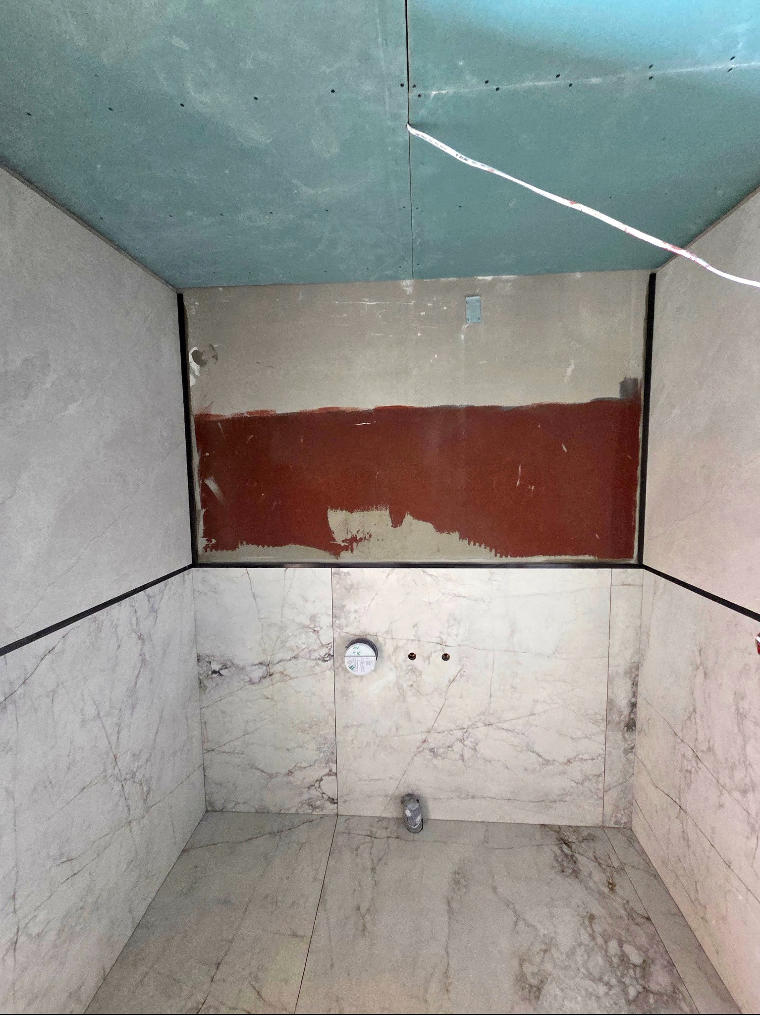 Wnętrze kabiny prysznicowej w trakcie wykańczania. Widoczne marmurowe płytki na ścianach i podłodze, surowy fragment ściany z instalacją i płyty gipsowo-kartonowe na suficie.