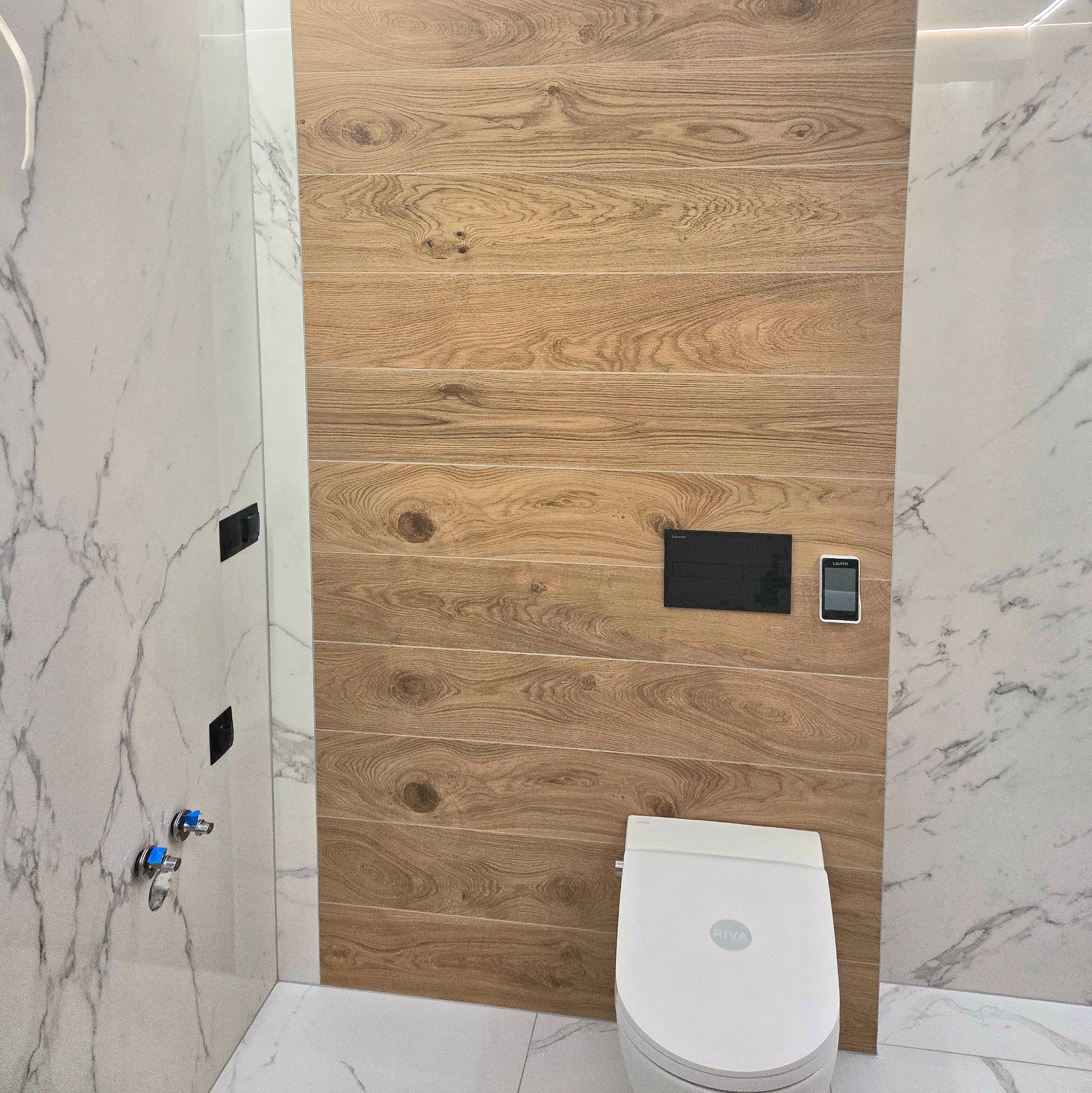 Nowoczesna łazienka z drewnopodobnymi płytkami na ścianie, marmurowymi płytkami i podwieszaną toaletą. Minimalistyczny design, eleganckie wykończenie.