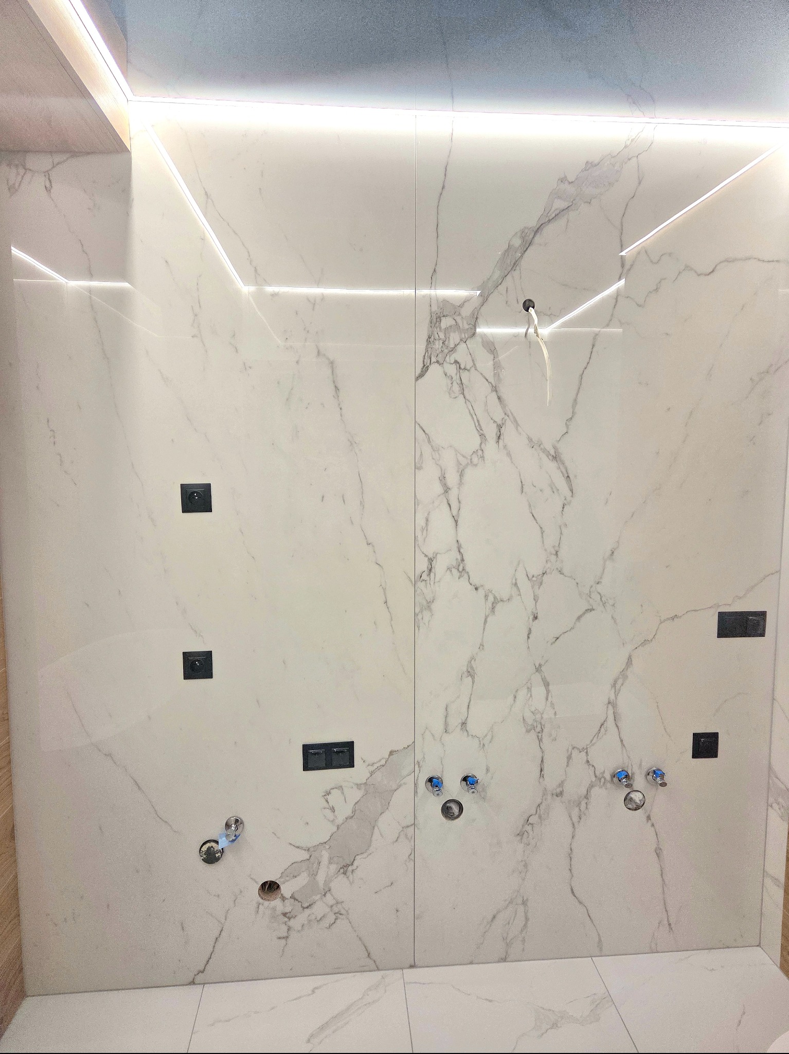 Wykończona ściana w łazience z białych płytek imitujących marmur, z zamontowanymi włącznikami i wyprowadzeniami wodnymi. Oświetlenie LED wzdłuż krawędzi.